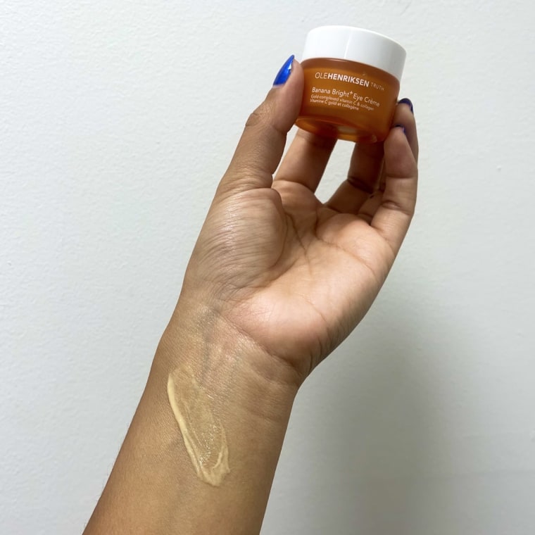 A woman holds Olehenriksen Banana Bright+ Eye Crème in her hand, and a swatch of the yellow eye cream is on her arm.