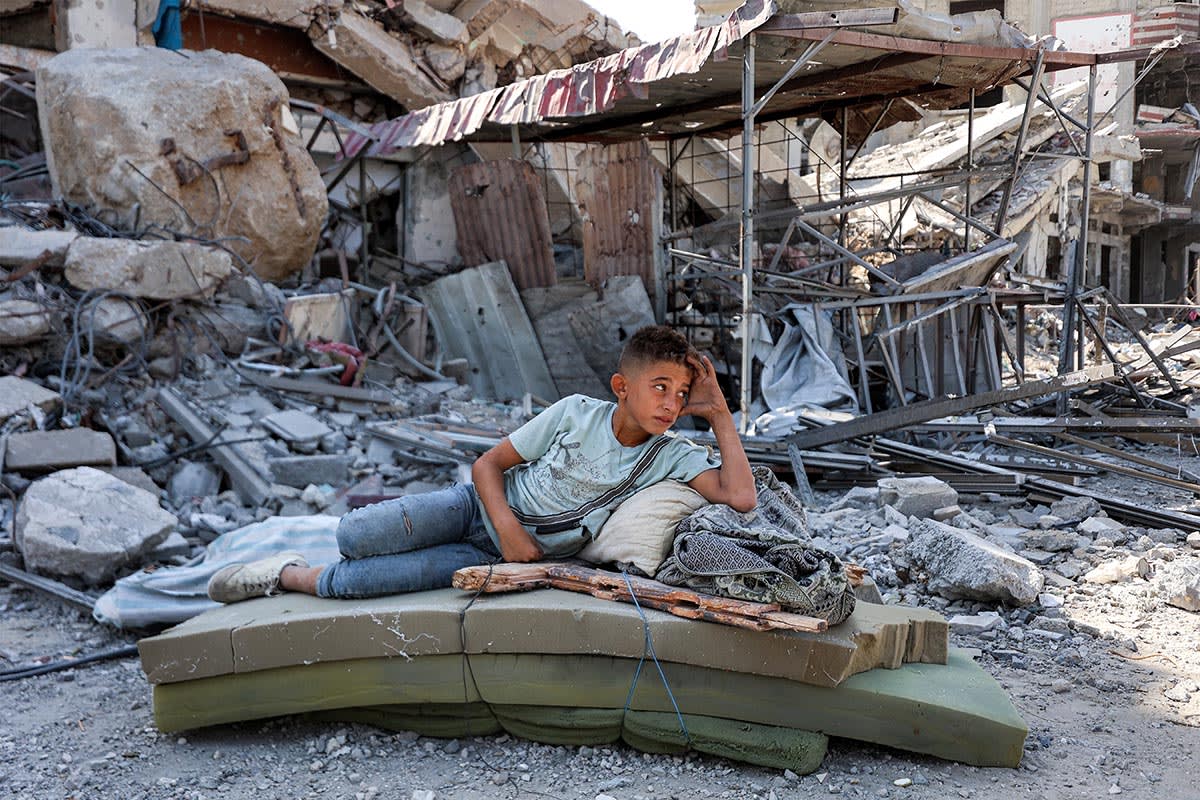 A boy lies on several foam mattresses by rubble outside a destroyed building in the center of Khan Younis