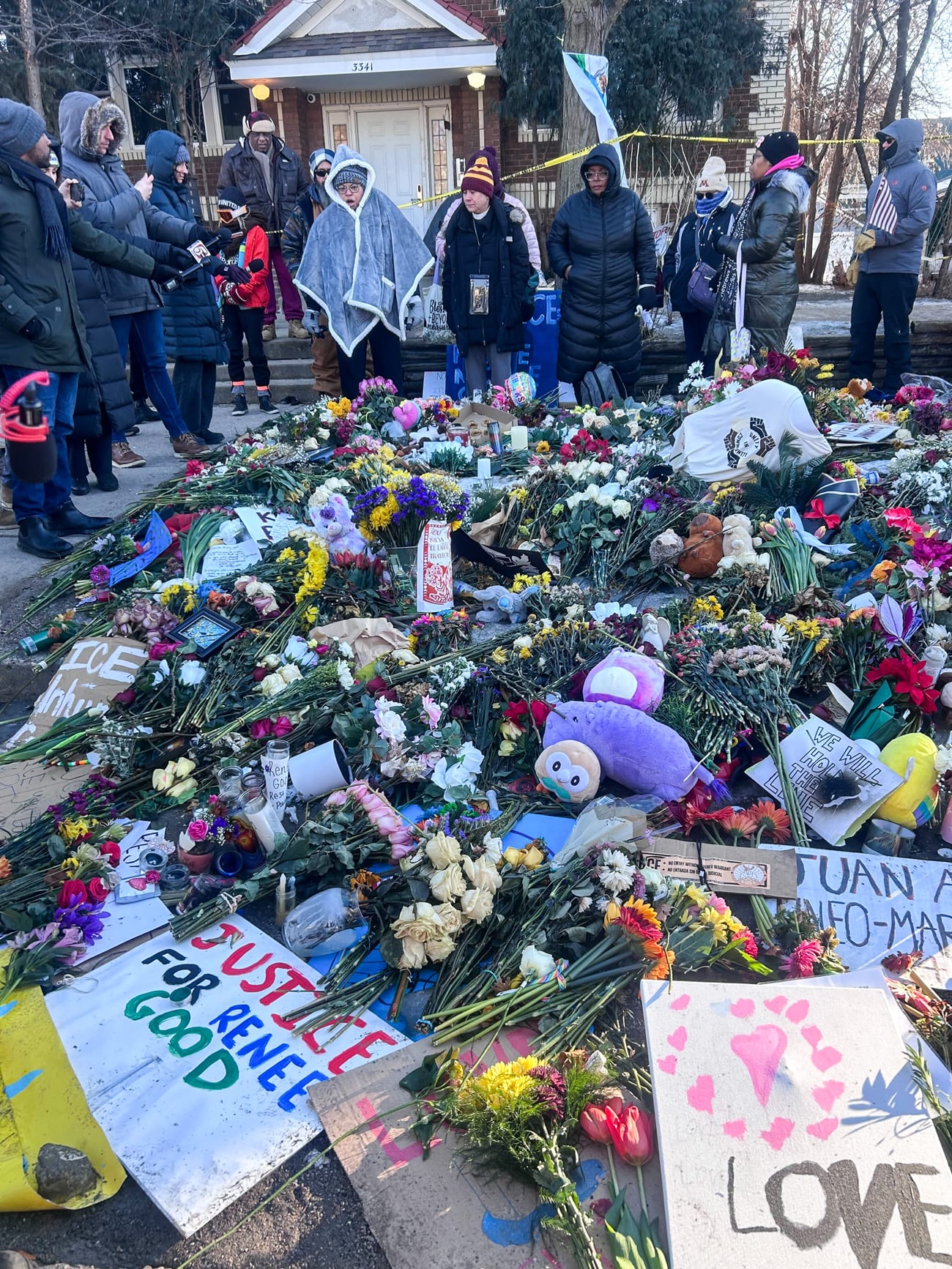 Flowers, signs, and stuffed animals on the street, people surrounding