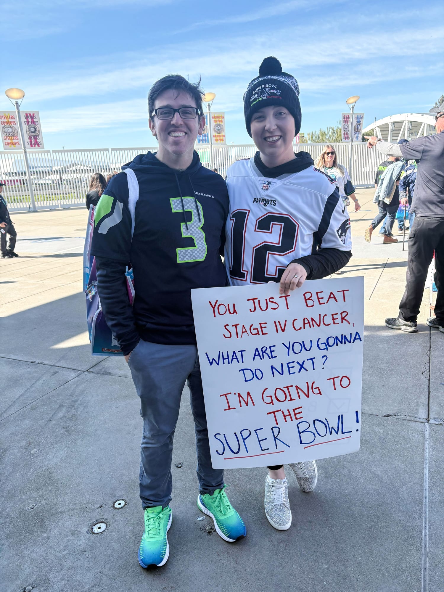 A man wearing a Seahawks jersey and woman wearing a Patriots jersey pose for a portrait, with a sign that reads: "You just beat stage IV cancer, what are you gonna do next? I'm going to the Super Bowl!"