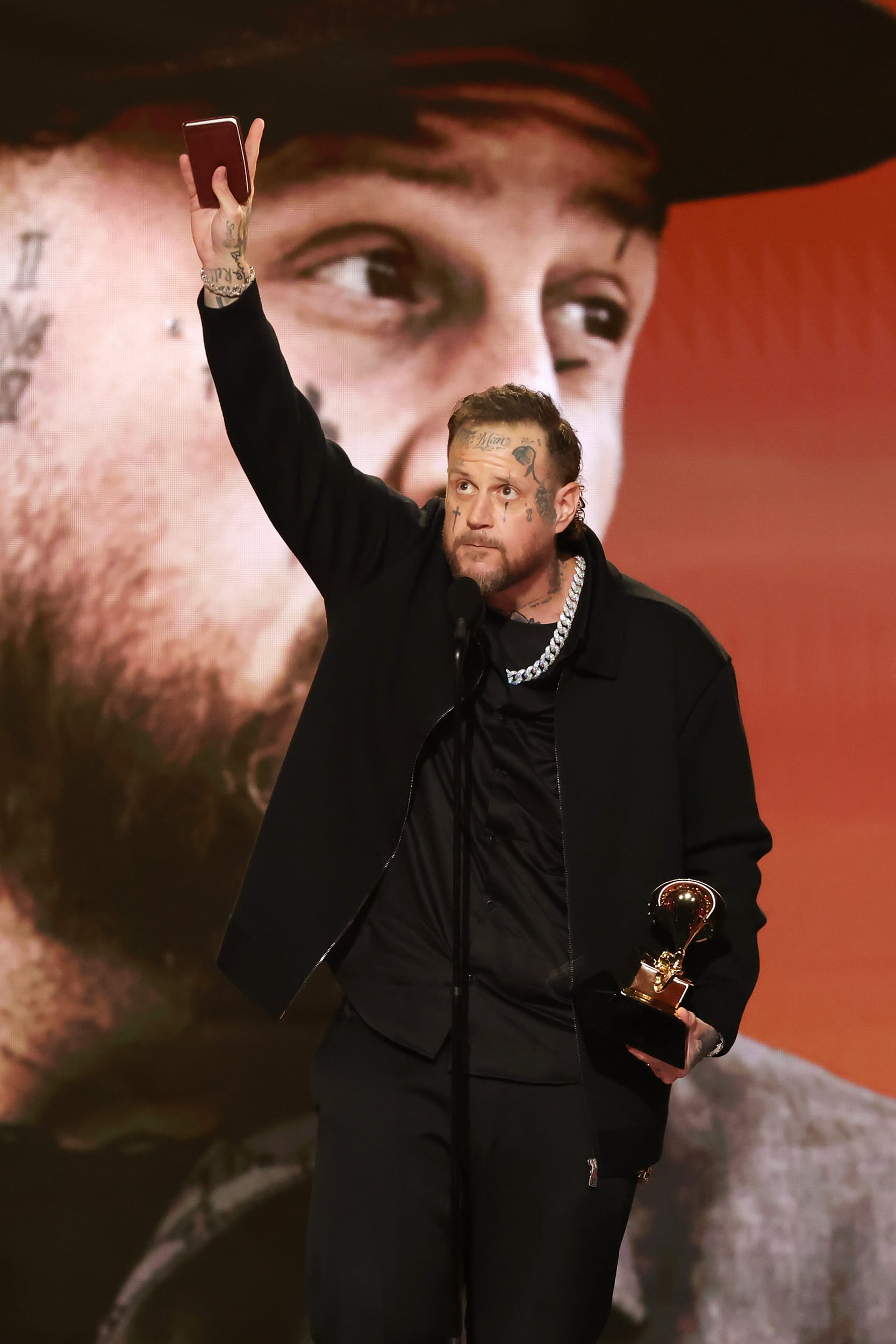 Jelly Roll holds a notebook up in one hand while holding a Grammy award in his other hand. He's standing at a microphone.