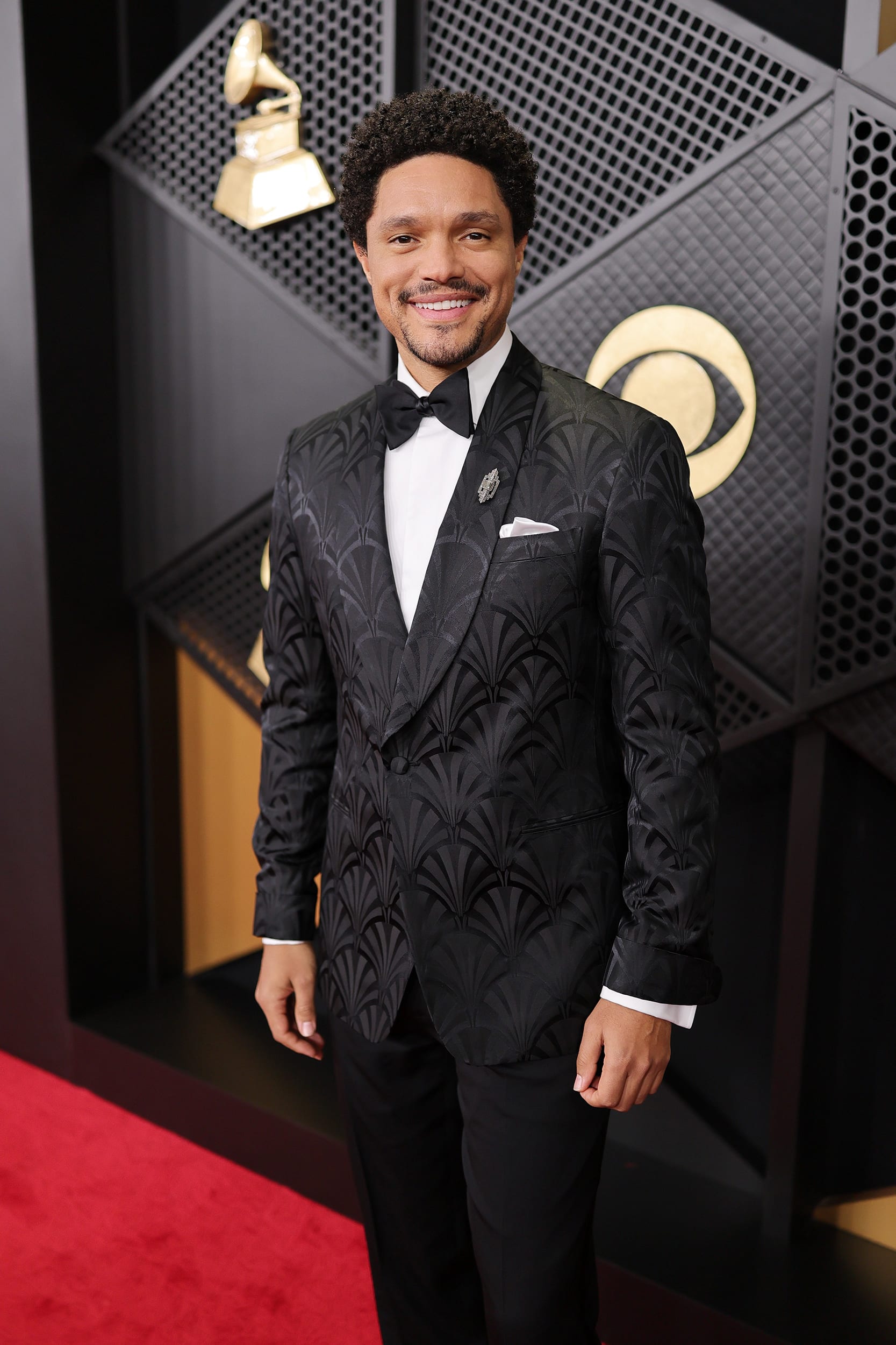 Trevor Noah smiles in front of a step-and-repeat backdrop, wearing a tuxedo.