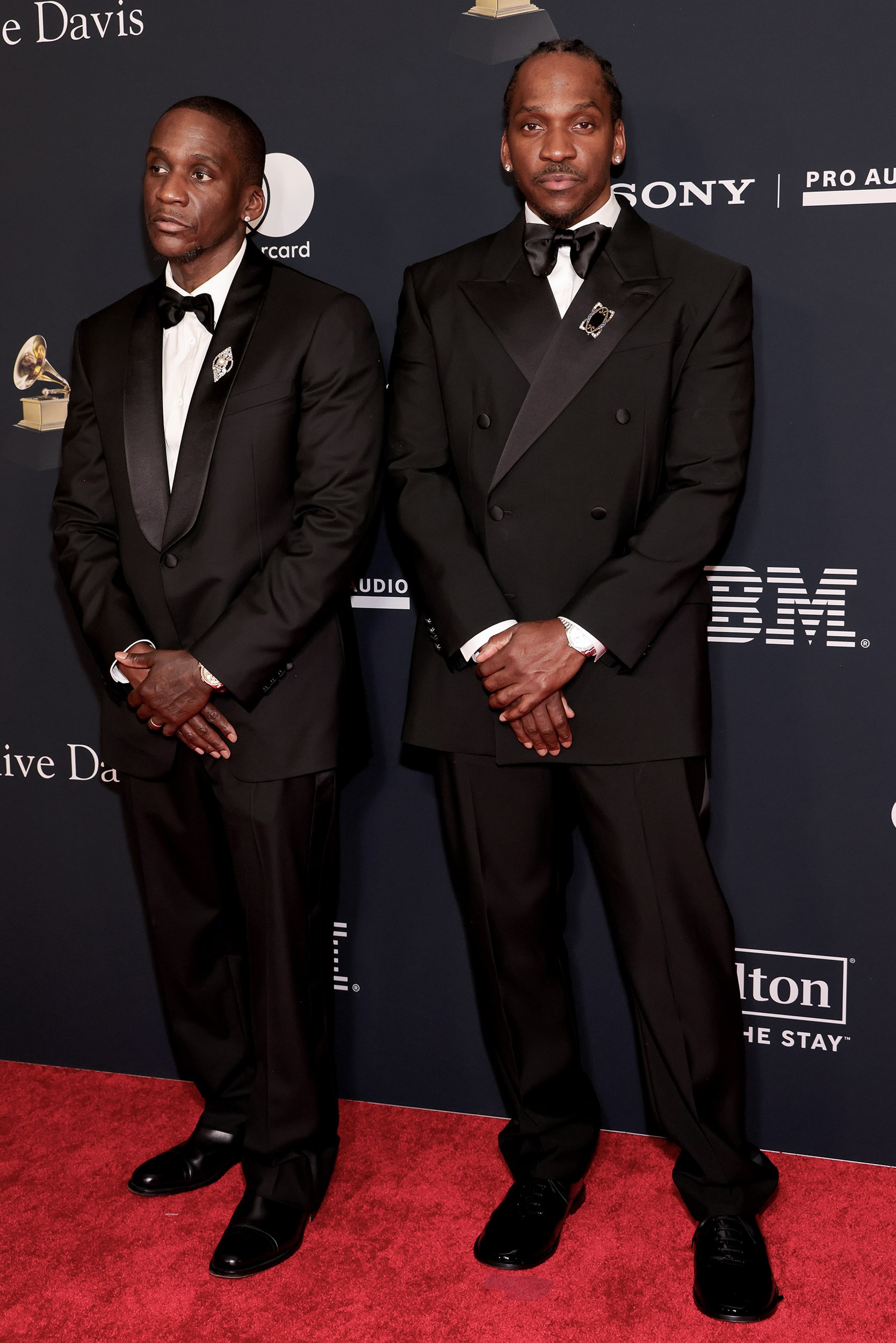 Malice and Pusha T pose against a step-and-repeate backdrop during the Grammys. They are both wearing black tuxedos.