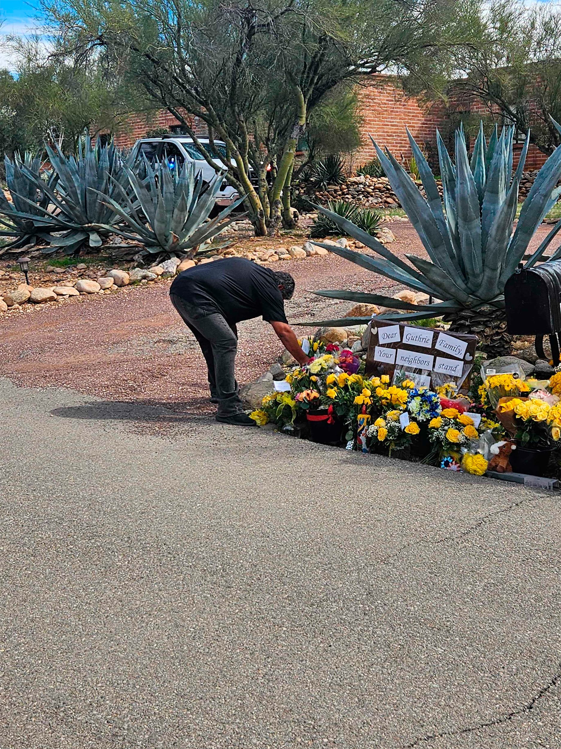 A man wearing black jeans and a black t-shirt bends over to place flowers in a pile in front of a home in the desert.