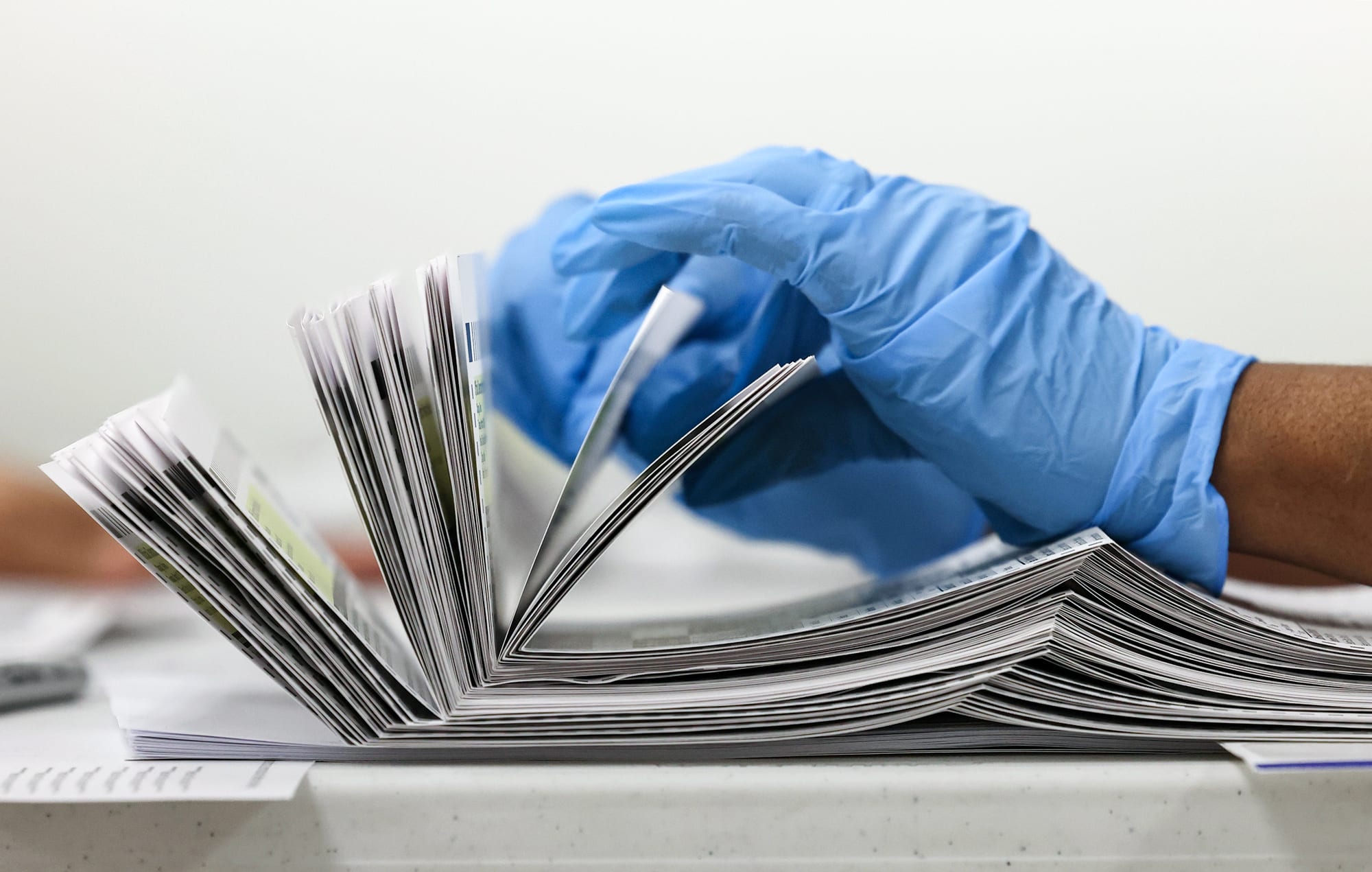 A gloved hand (blue) counts a stack of mail-in ballots on a table, side view