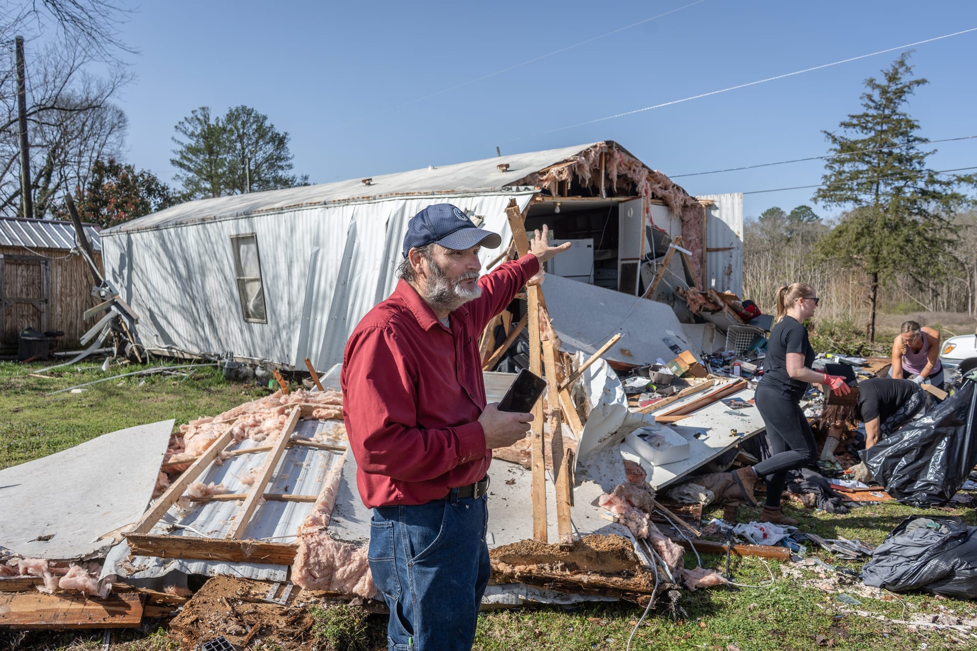 A person points towards a damaged house outside while standing among debris on the front lawn