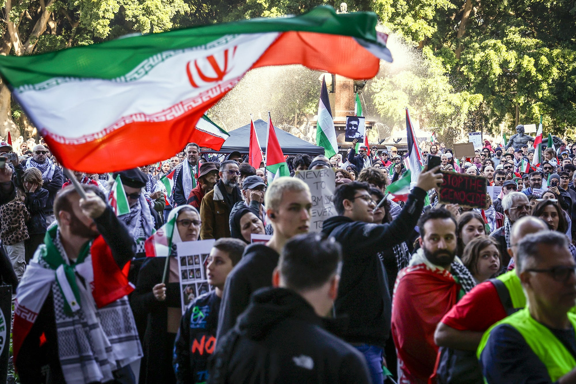 A large crowd gathers to protest, signs and Iranian flags are held