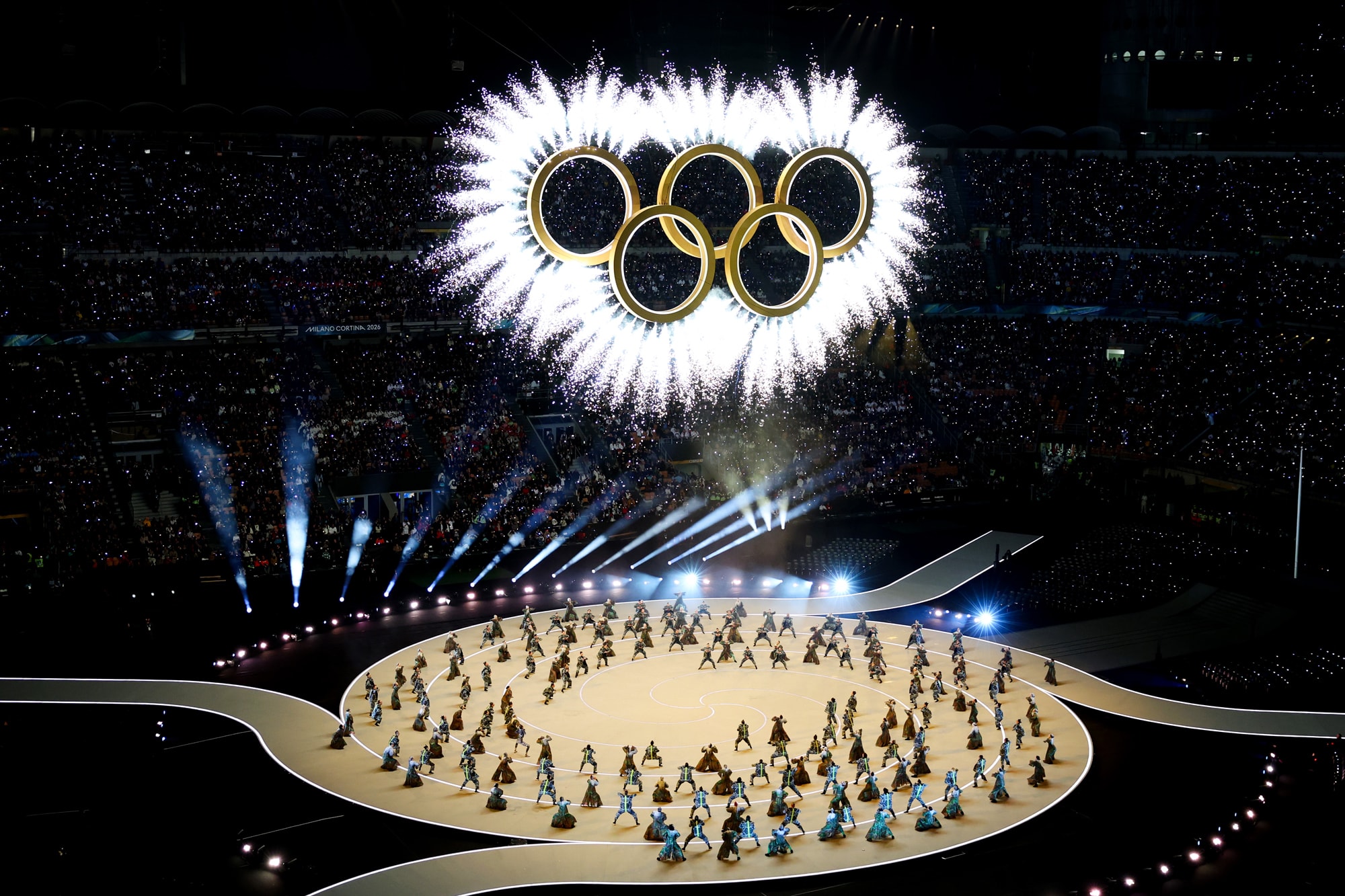 Dancers perform on stage, Olympic rings with fireworks are seen above