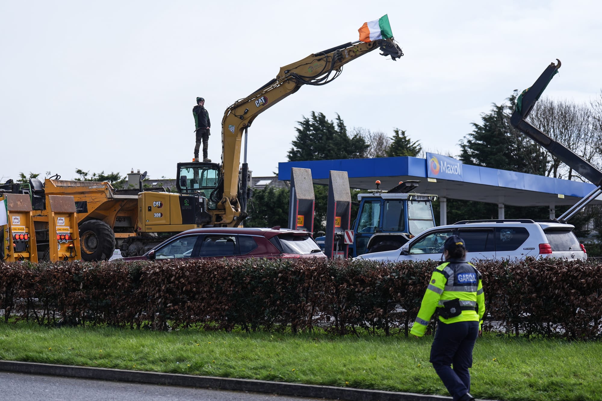 A person stands on an excavator vehicle that is blocking street traffic, an Ireland flag is on the top of the vehicle