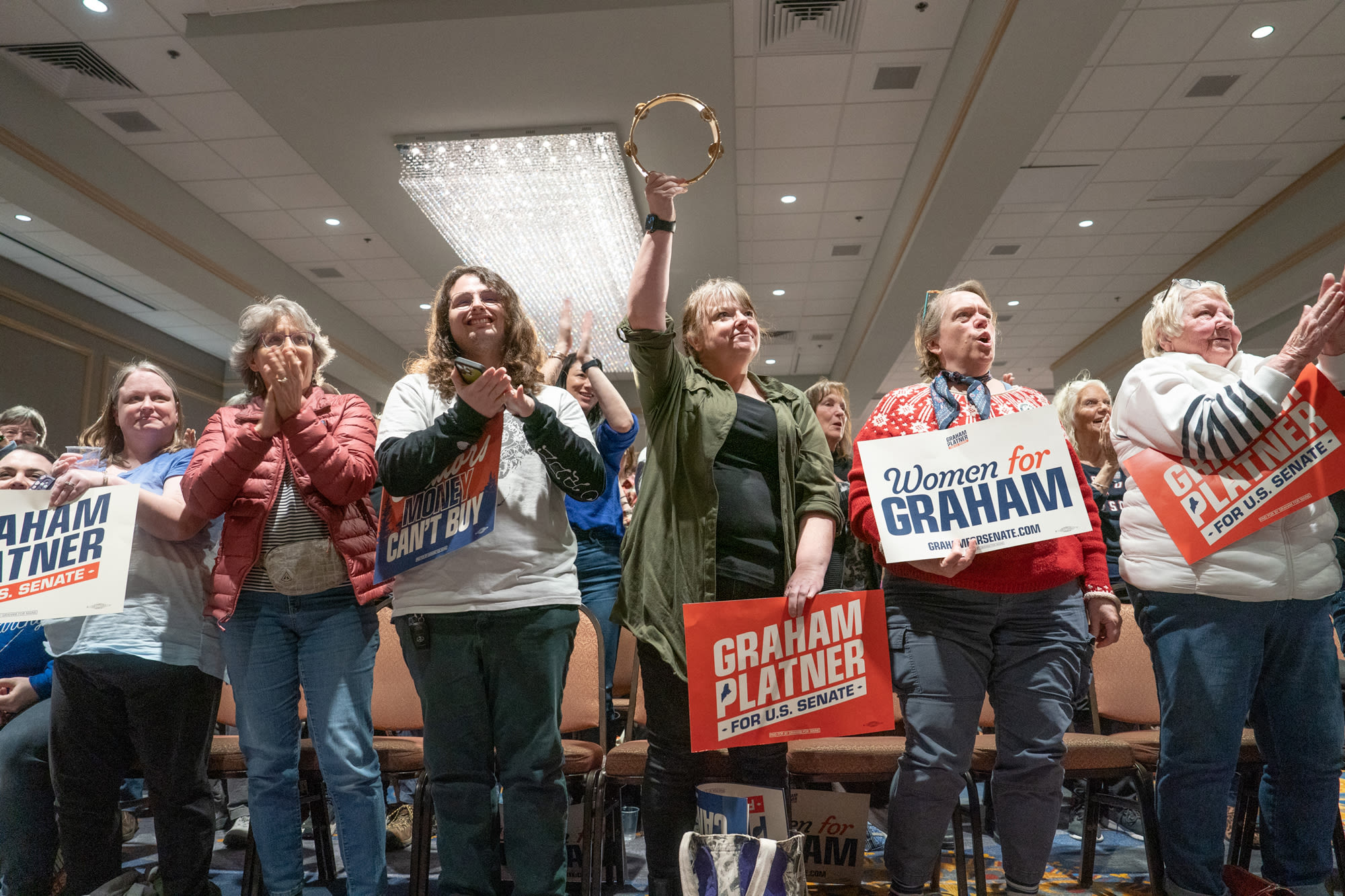 Supporters cheer and holds signs (and a tambourine) inside of an event hall