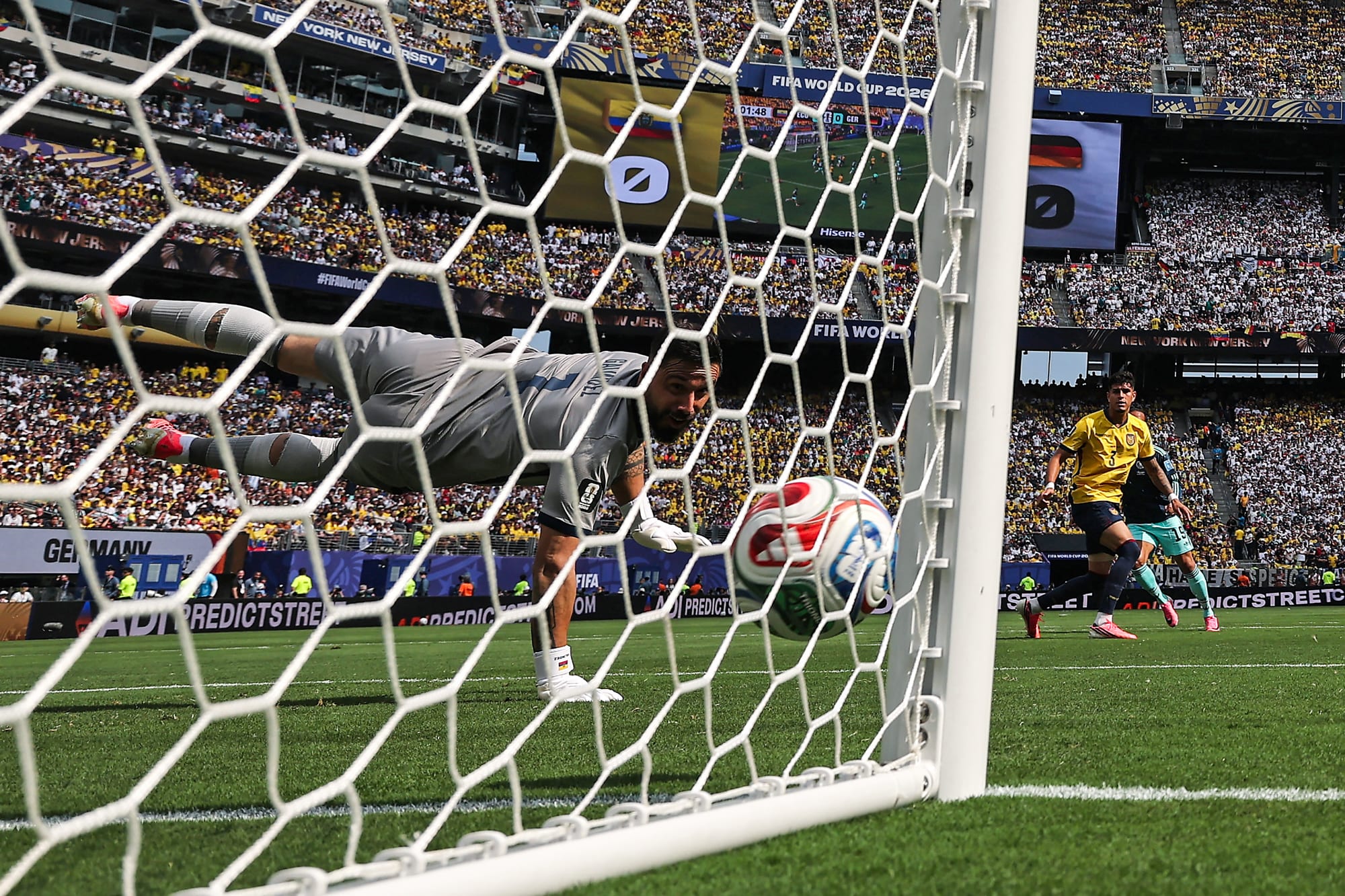 A ball is kicked into a net and a goalie dives to stop it on the soccer field, as seen from behind the net