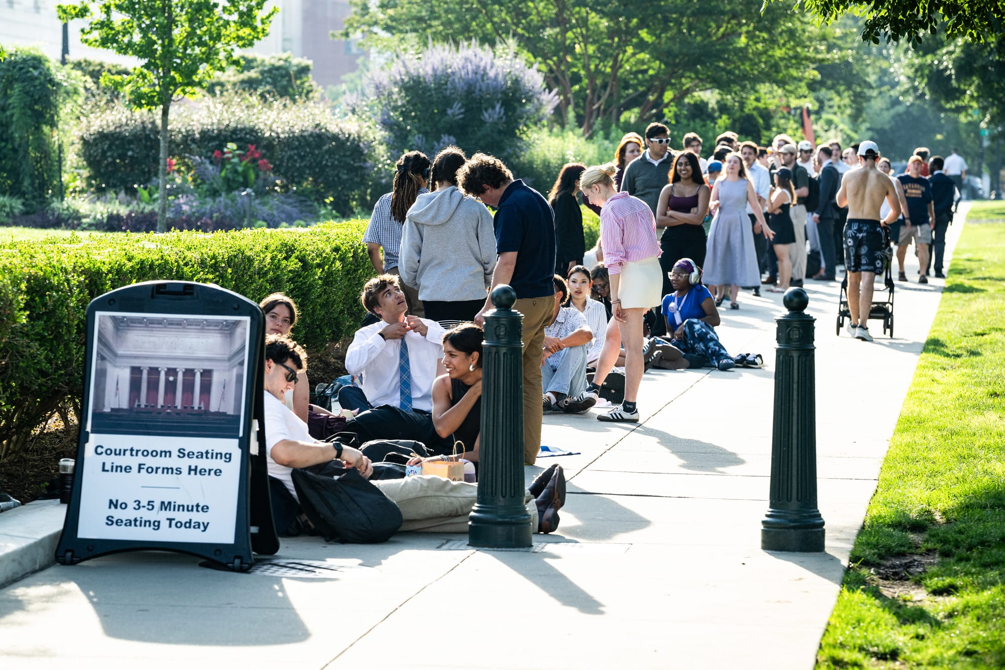 People wait in line on a sidewalk, some people seated on the ground, a sign in front of the line reads "courtroom seating line forms here, no 3-5 minute seating today" with a picture of a courtroom on top