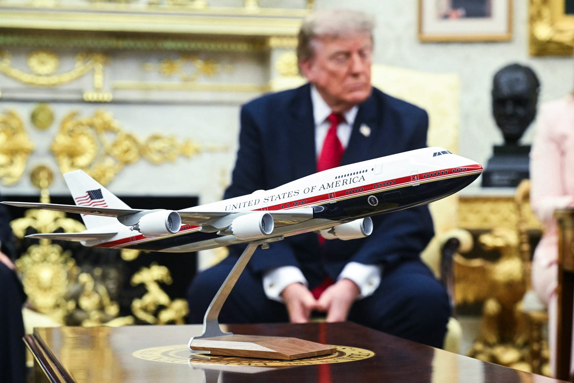 An Air Force One model sits on a table in the Oval Office, a blurred Donald Trump can be seen sitting on a chair behind it