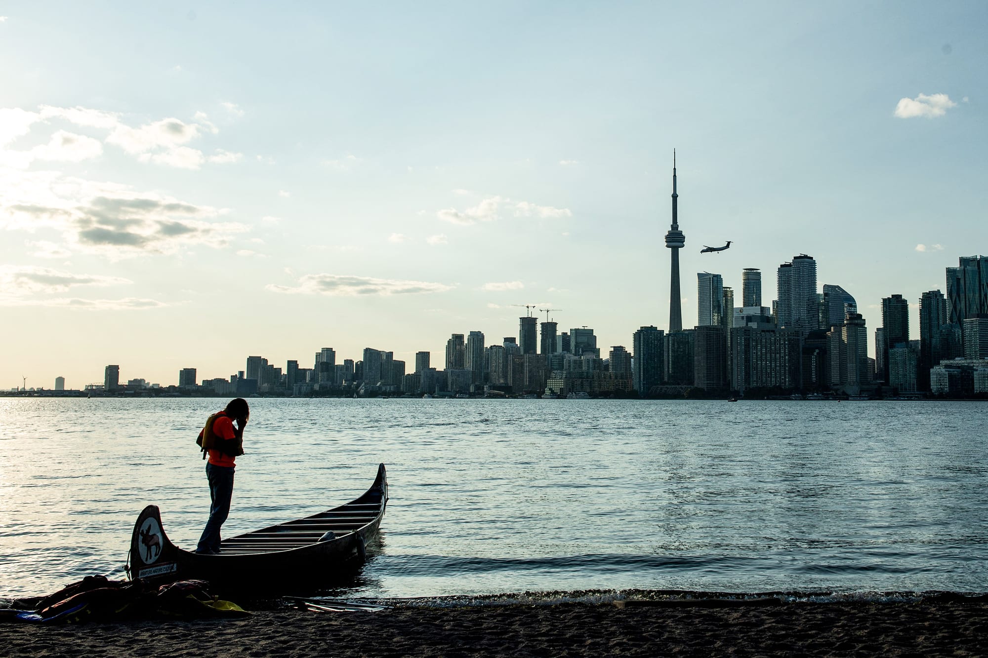 A person stands by a canoe resting on the calm water of Lake Ontario in front of the CN tower (Canadian National) and the Toronto skyline