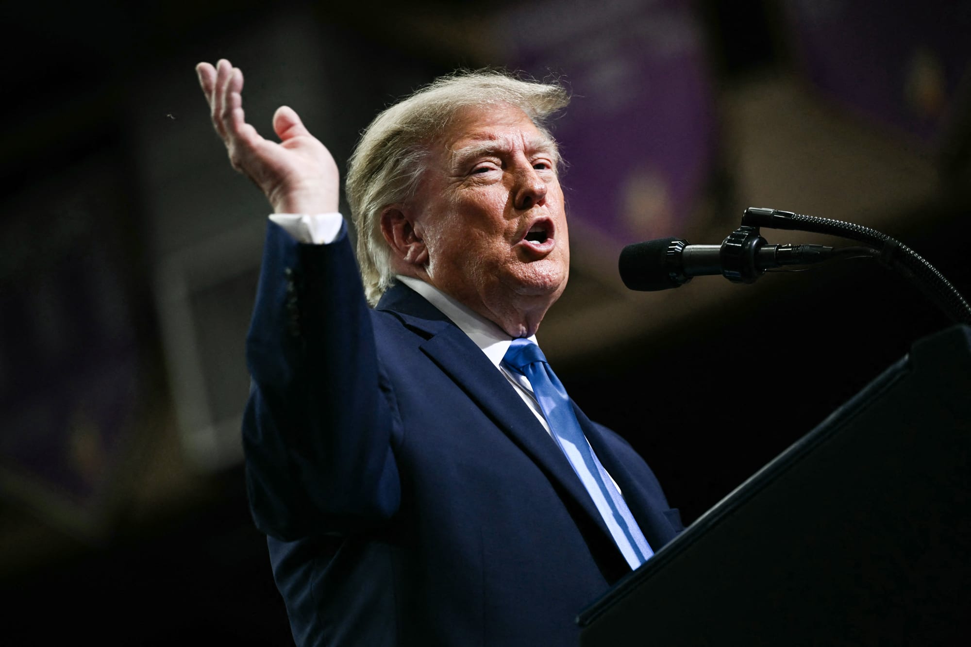 Donald Trump speaks at a podium, lifts one hand up in the air