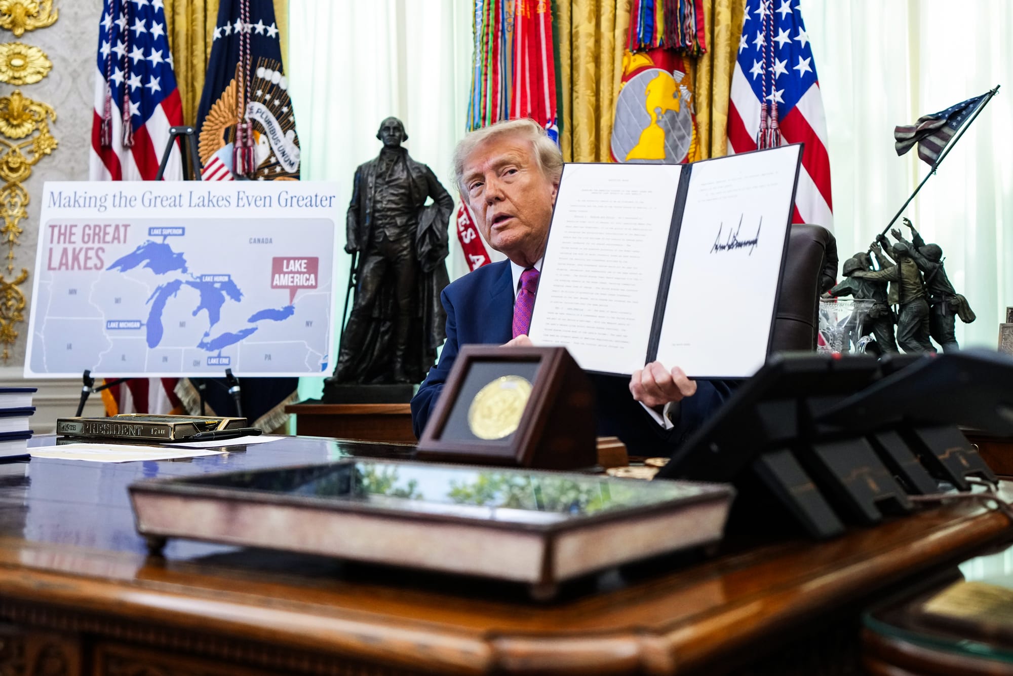 President Donald Trump displays a signed executive order while seated at his desk in the Oval Office, next to him is a poster showing a map of the great lakes that reads "Making the Great Lakes even greater" with Lake Ontario labeled as Lake America