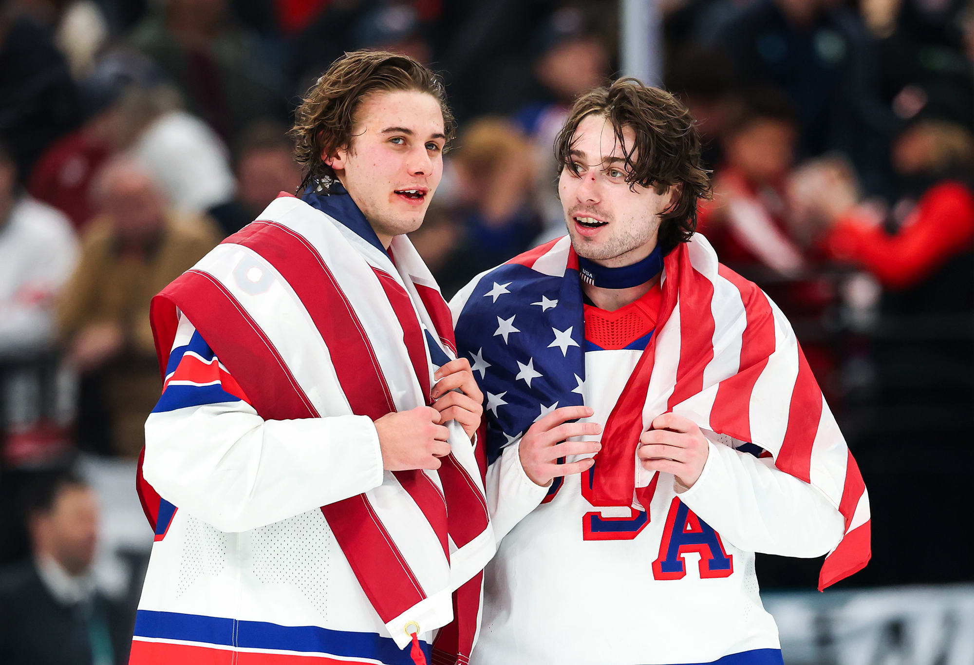 Jack Hughes, left, and Quinn Hughes speak with American flags draped over their shoulders on the ice