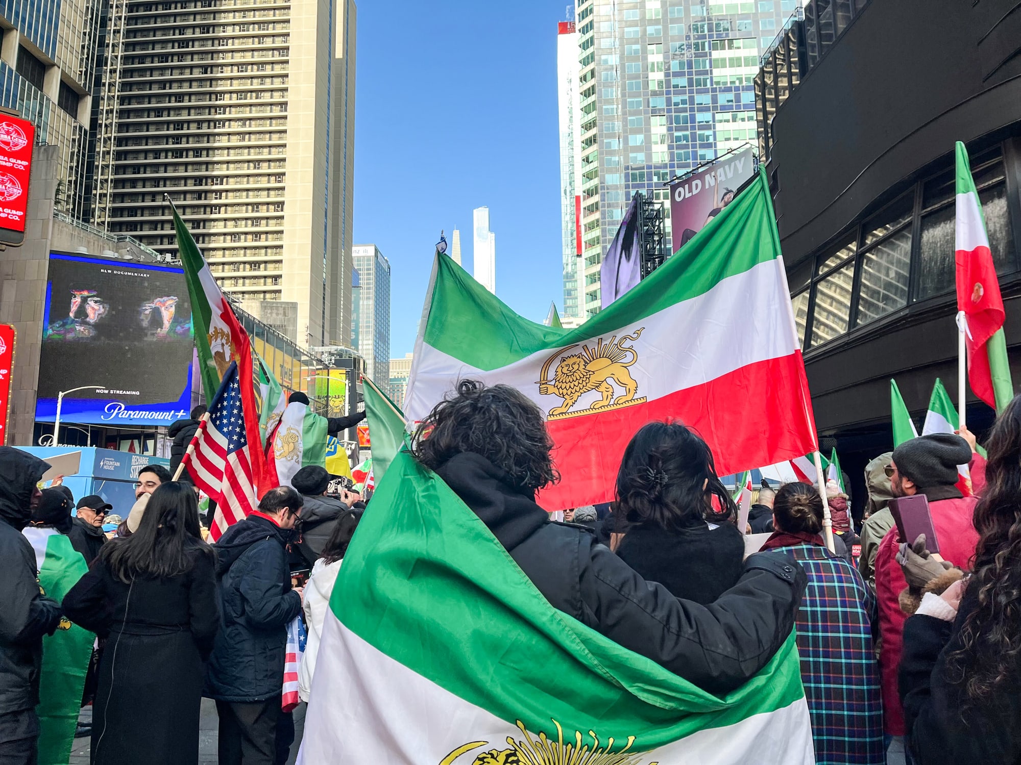 A crowd gathers outside with Iranian, Israeli, and American flags
