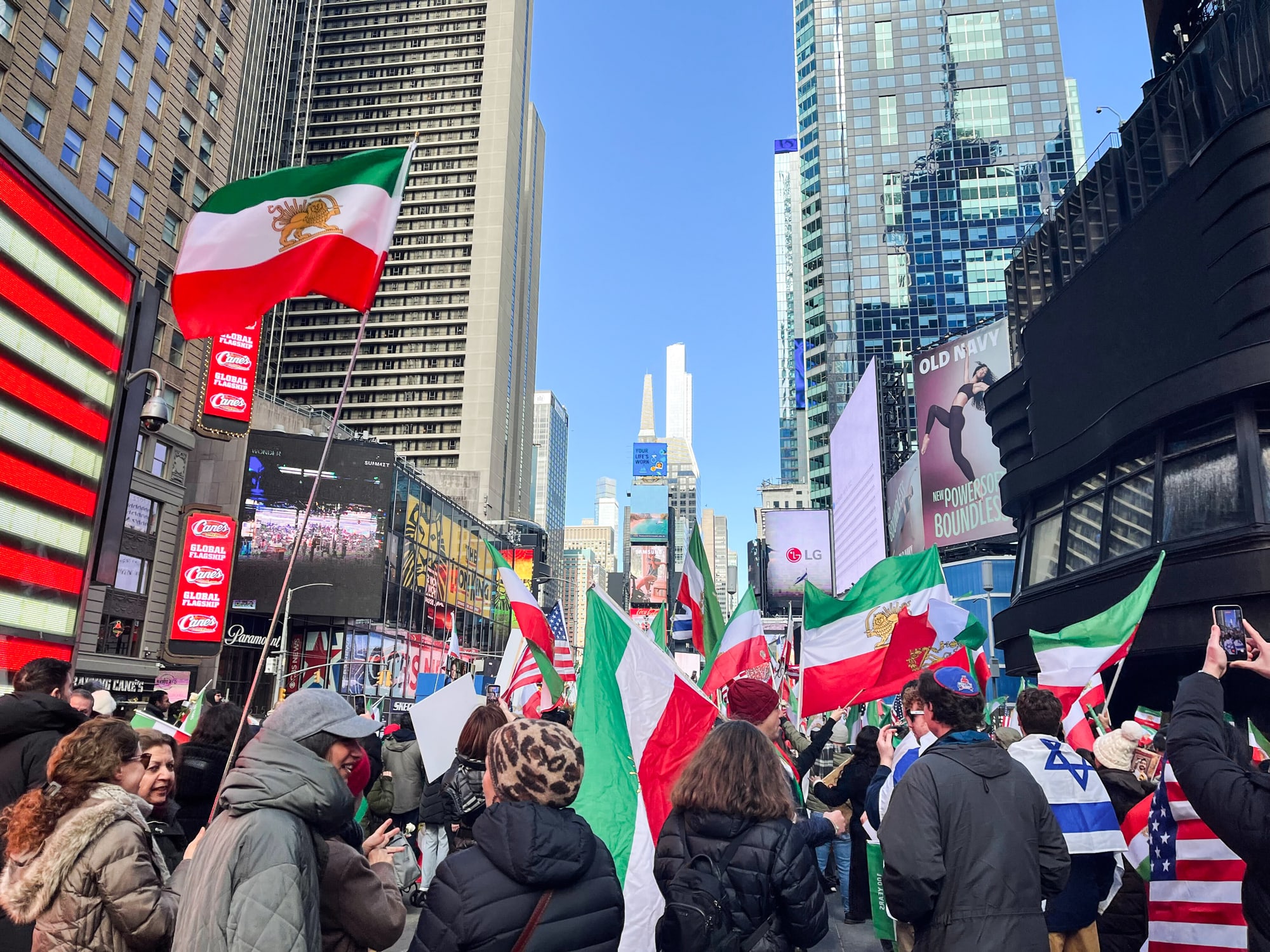 A crowd gathers outside with Iranian, Israeli, and American flags
