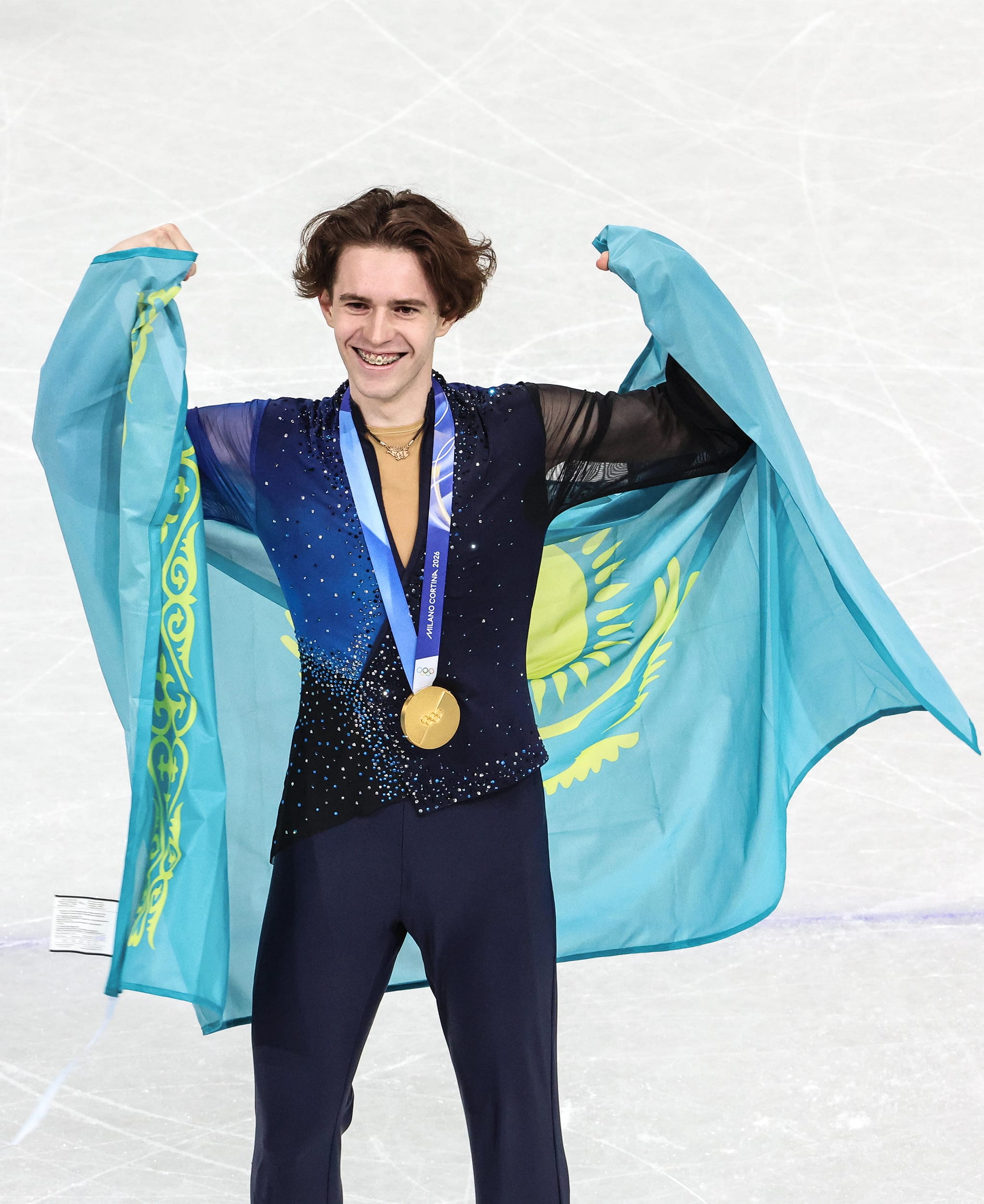 Mikhail Shaidorov holds the blue and yellow national flag of Kazakhstan around his shoulders as he smiles with a gold medal on his neck.