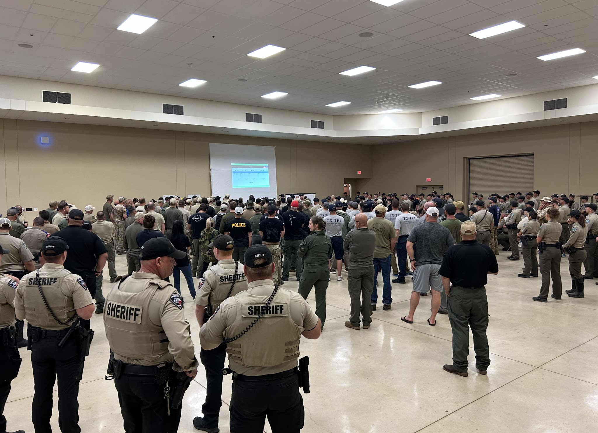 Sheriffs, police officers, and volunteers stand inside of a room, looking at a projected screen on the wall