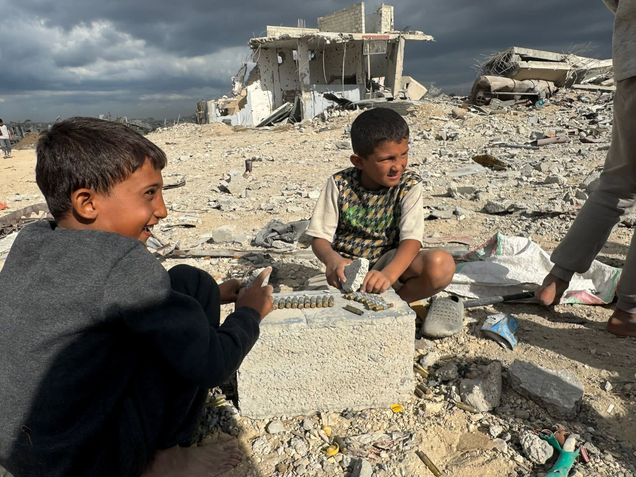 Children sit amongst rubble on the ground outside and make belts out of bullets