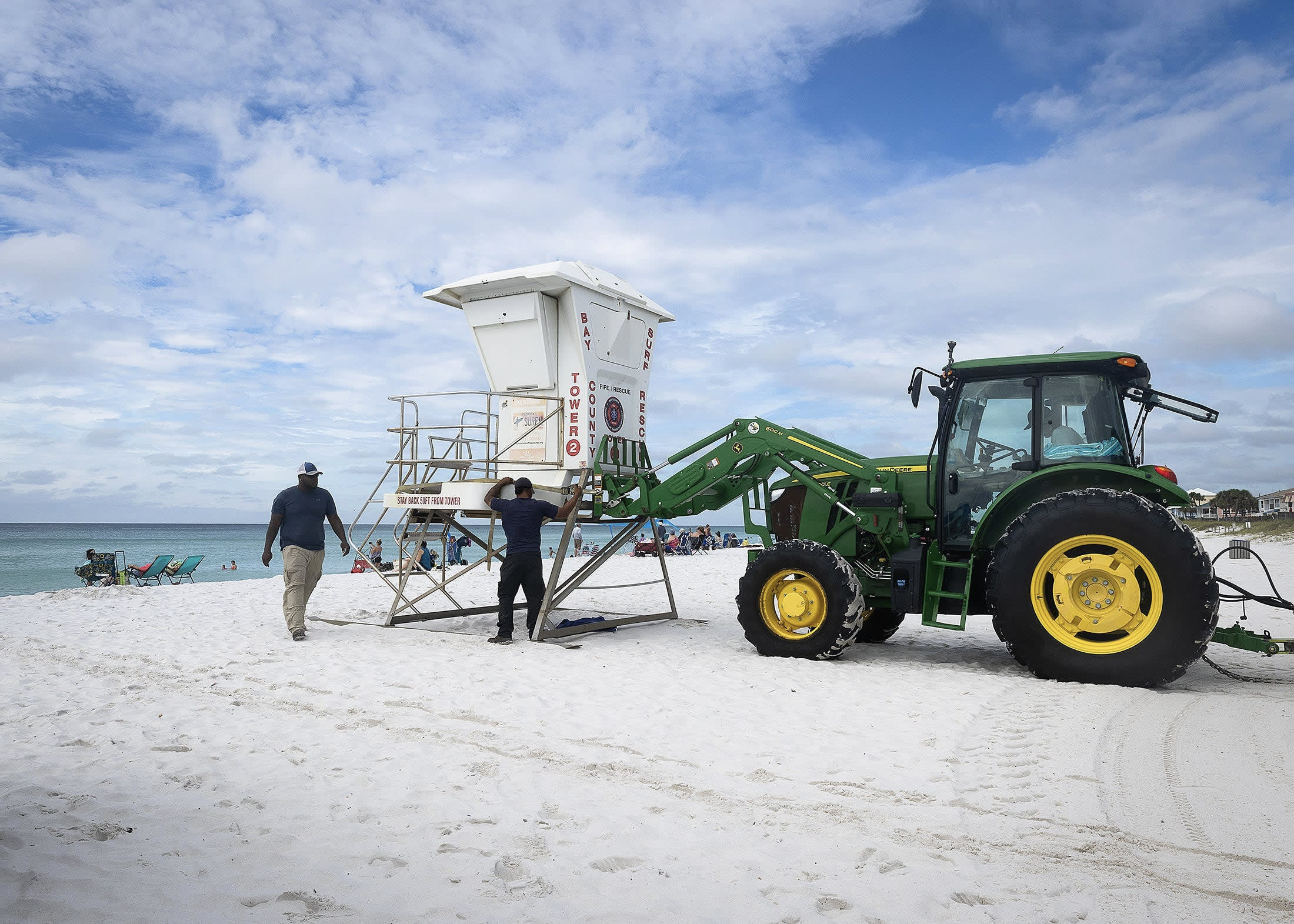 A front end loader lifting a white lifeguard tower on a Florida beach under cloudy skies.