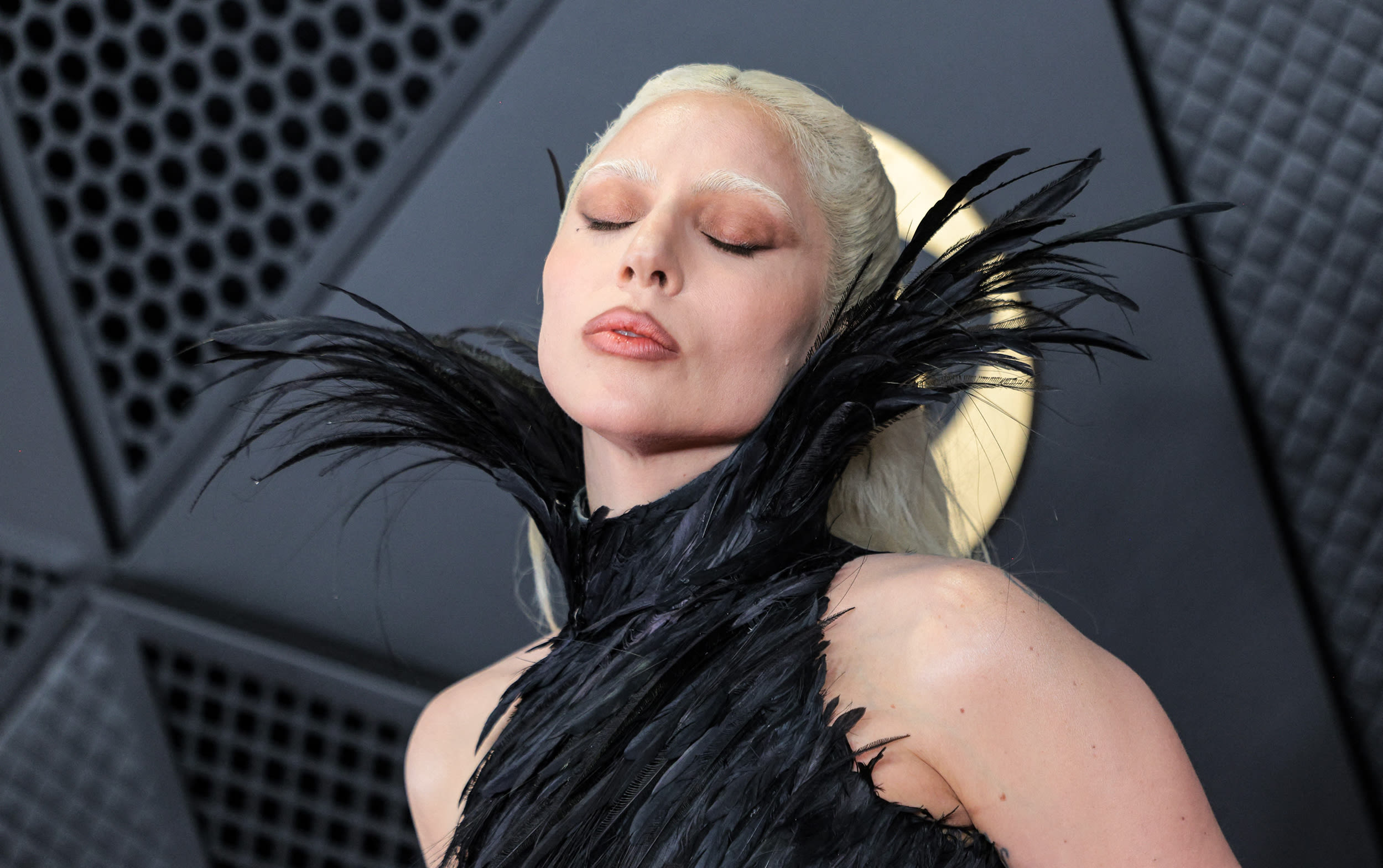 Lady Gaga, wearing a black feathered dress with a high collar, poses for a photo with her eyes closed.