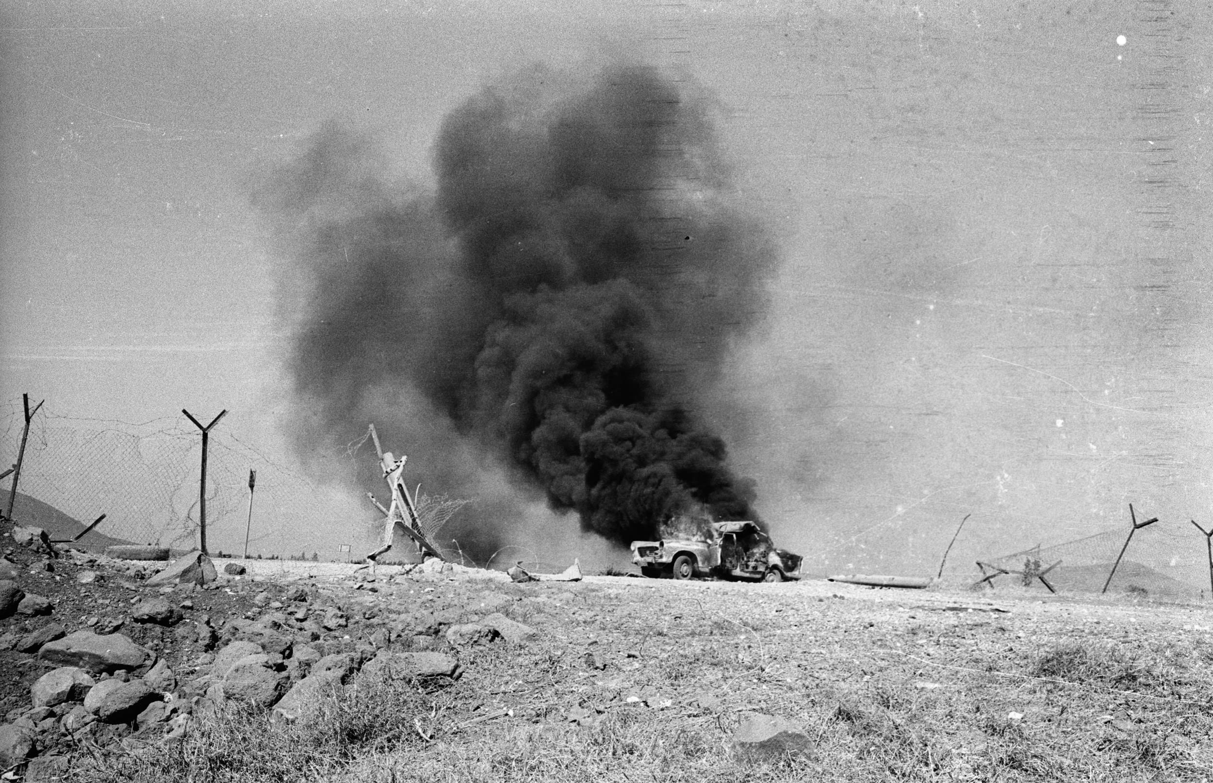 A black and white photo of smoke billowing from a bombed civilian car on a hillside in Golan Heights, Israel
