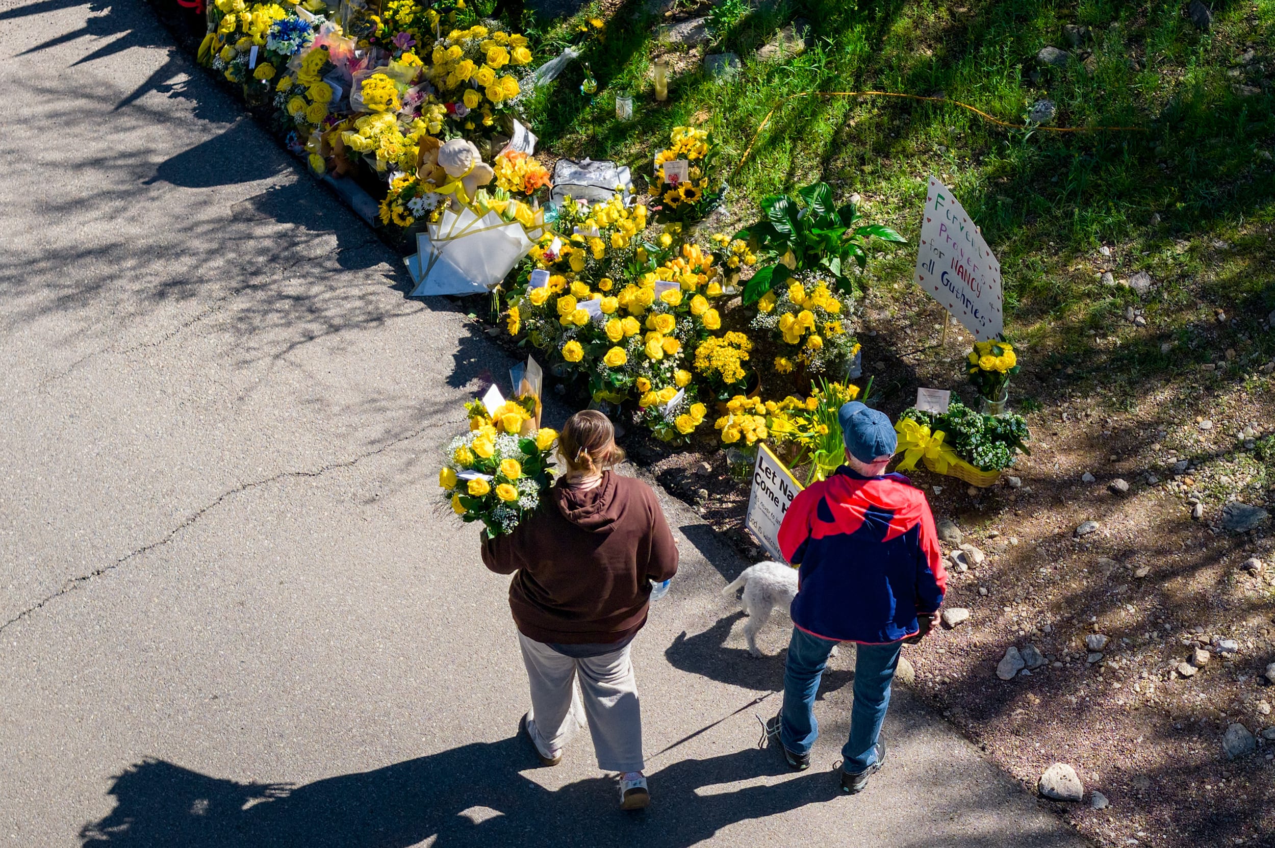 Two people are seen approaching a memorial of yellow flowers from an elevated perspective. One of them carries a bouquet of similar yellow flowers. 