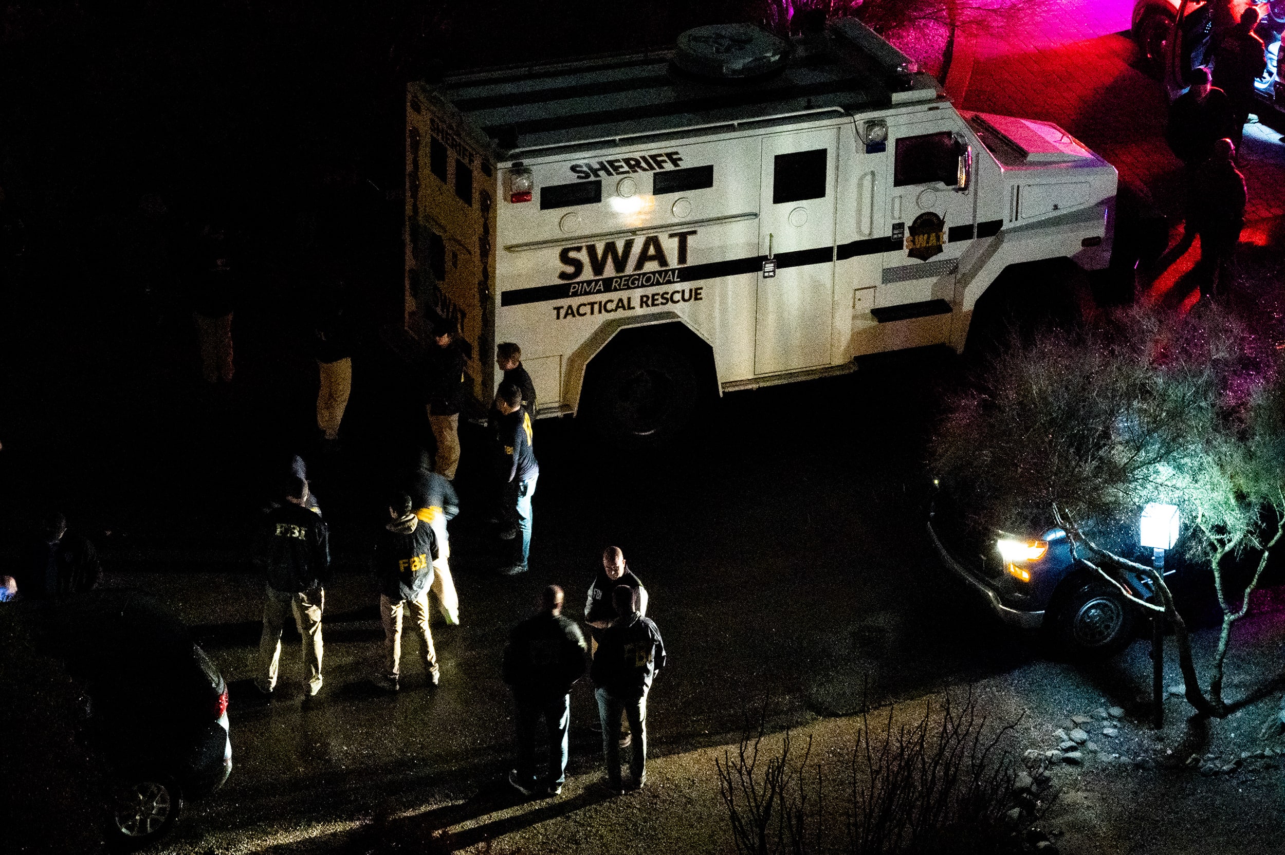 Law enforcement officials stand in front of a white SWAT van on a dark residential street.