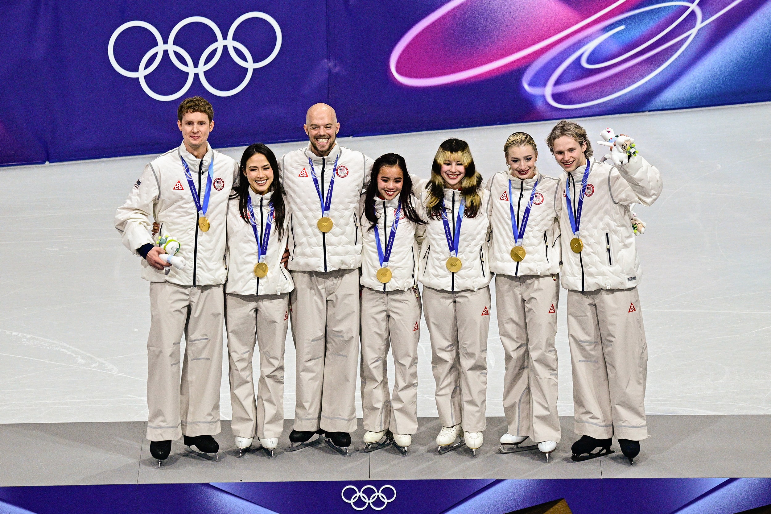 Seven olympic figure skaters in white warmup suits stand with gold medals around their necks.