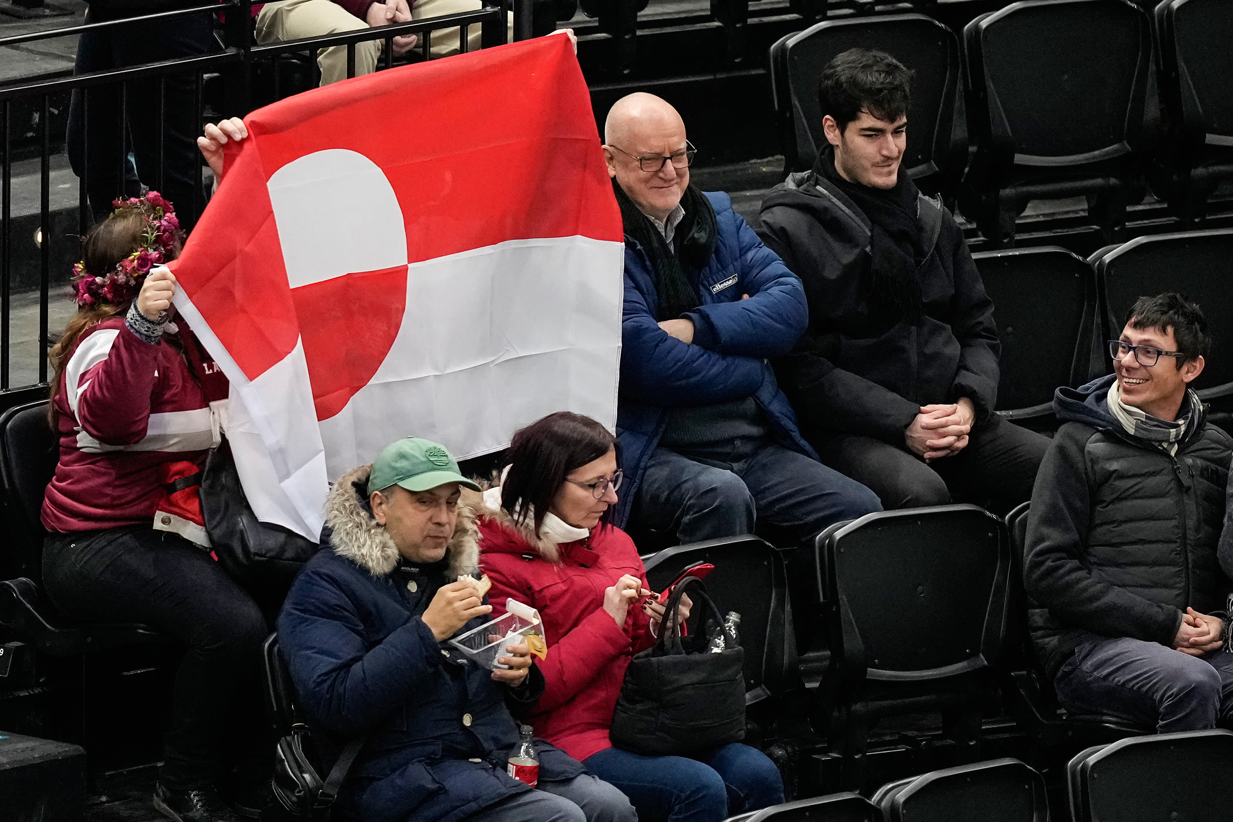 Two people hold up the red and white national flag of Greenland while seated in the stands at an indoor hockey stadium.