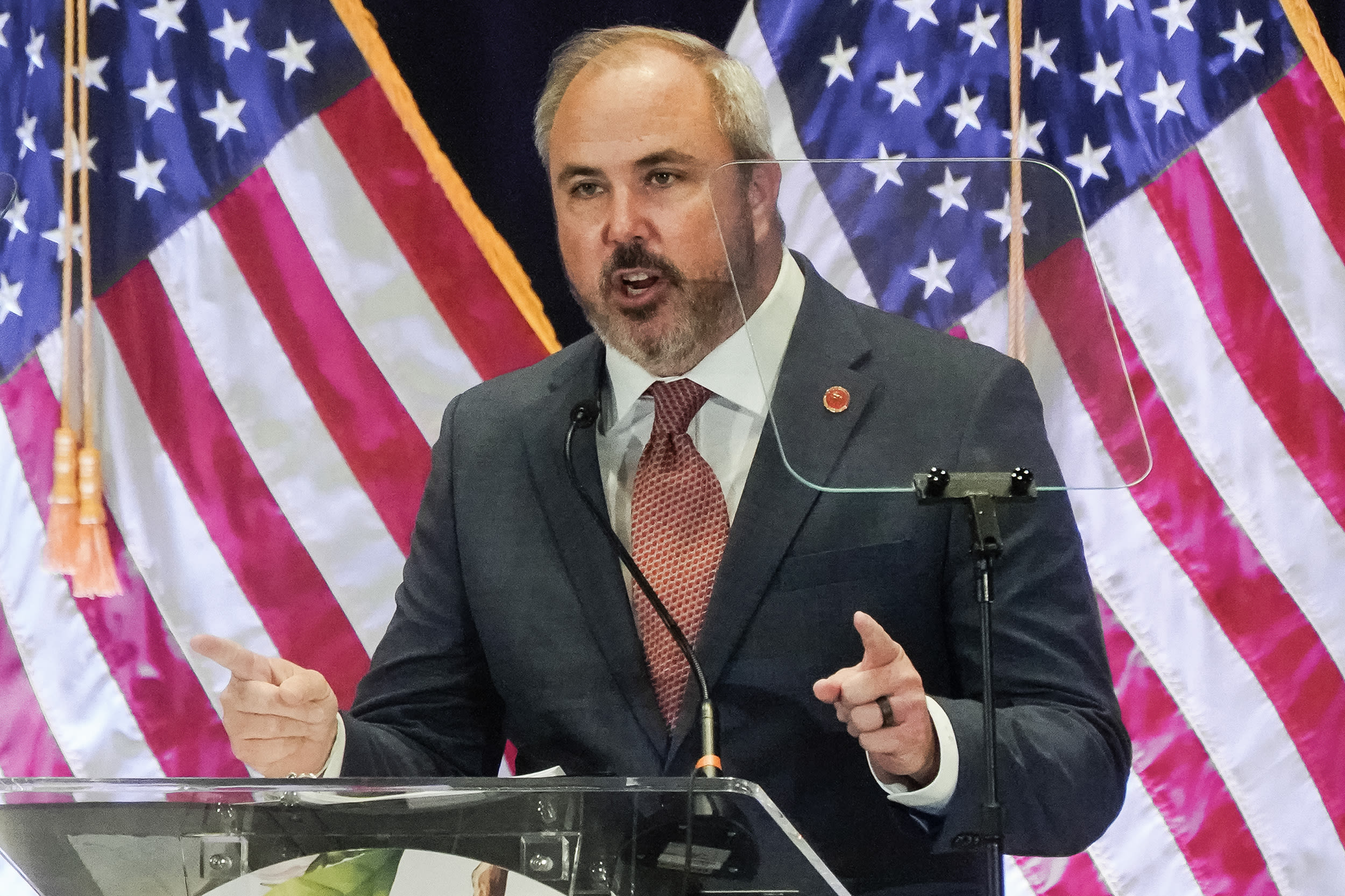 Joe Gruters gestures with both hands while standing at a clear lectern in front of two American flags.