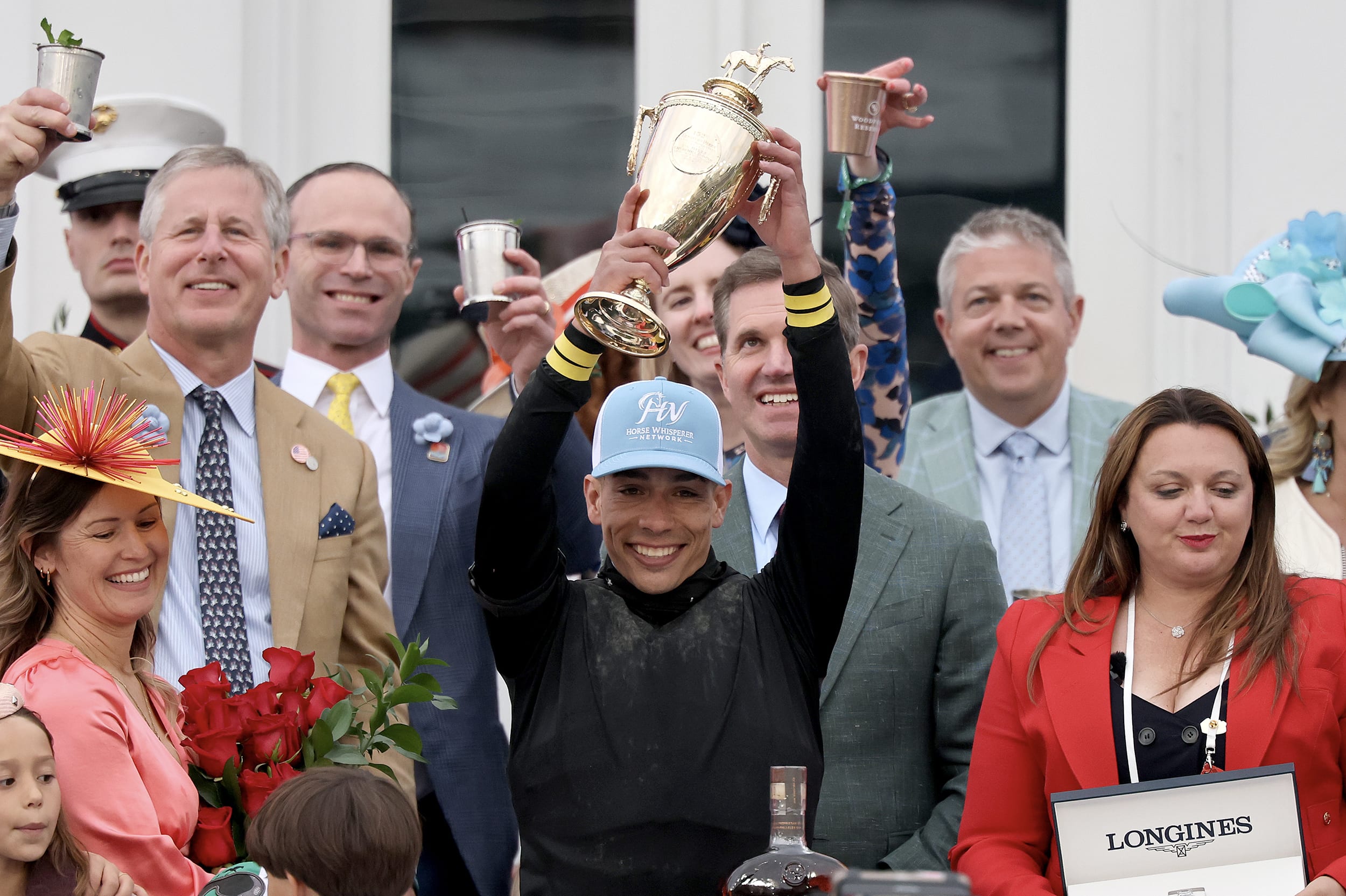 A jockey holds up a trophy while smiling broadly, surrounded by other celebrants.