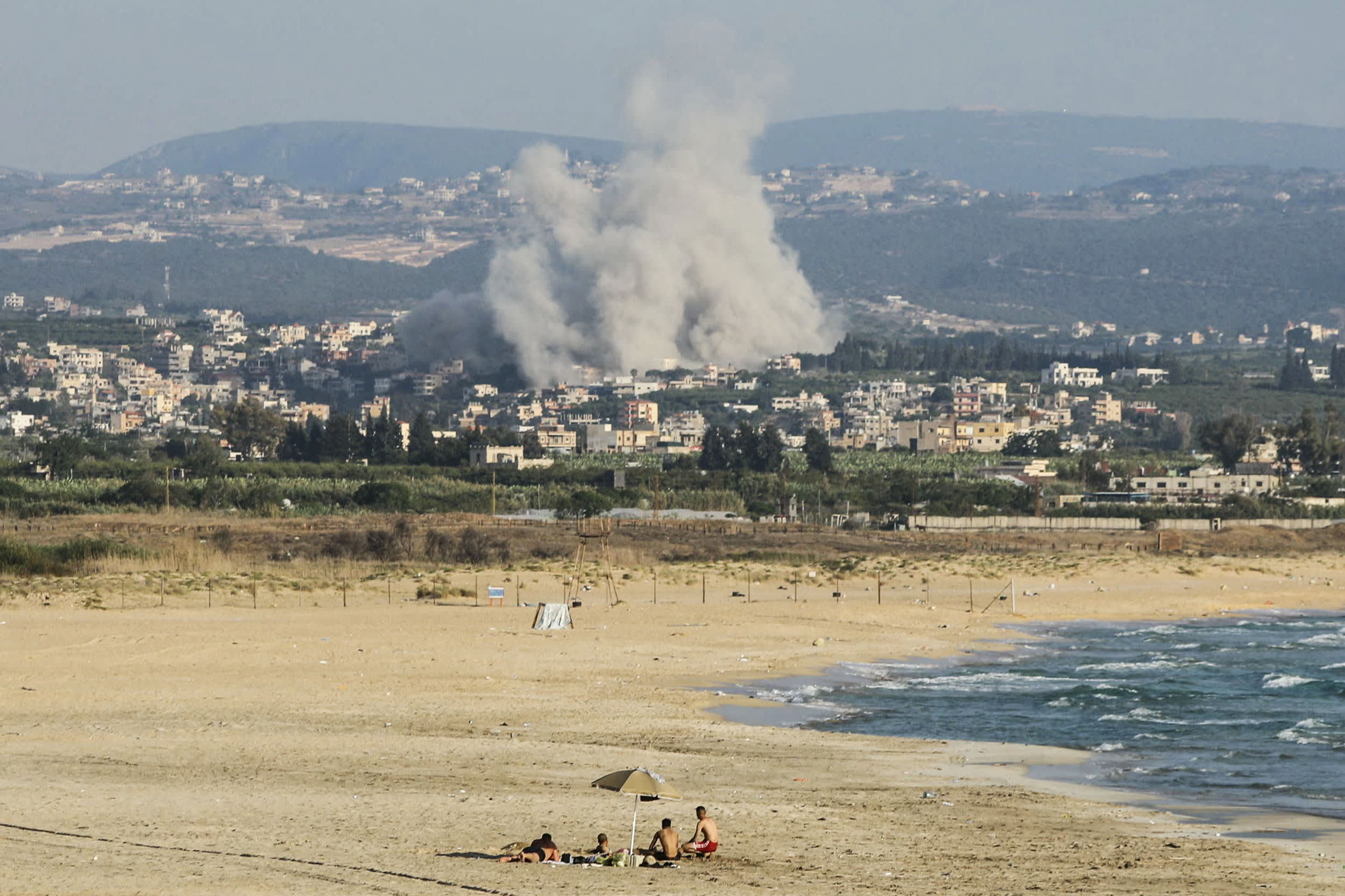 A photograph taken from the southern city of Tyre shows people sunbathing on the beach as smoke rises from the site of an Israeli airstrike in the village of Deir Qanoun Ras al-Ain on June 8, 2026.