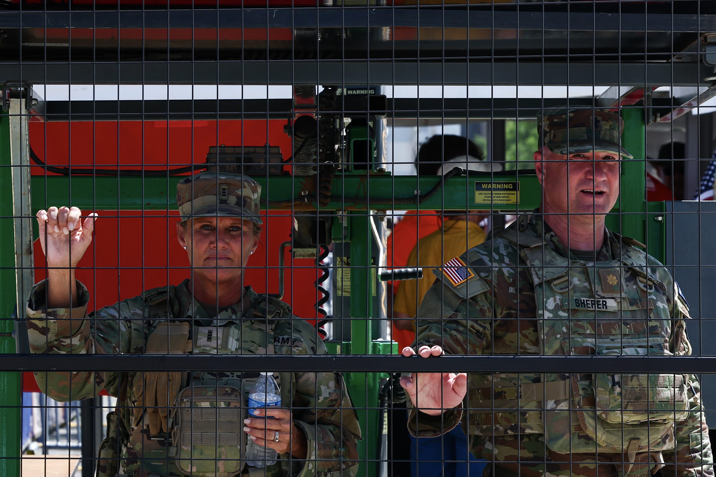 Two soldiers in camouflage sheltering from the sun behind a fence.