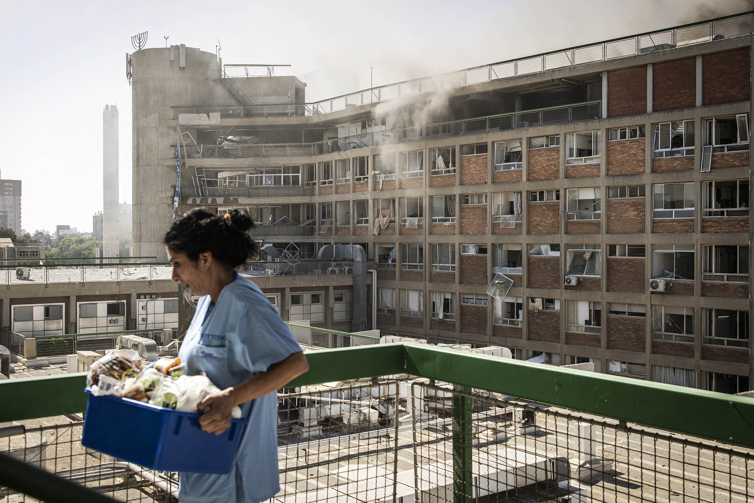 A nurse carries medical supplies past a building with smoke billowing out at Soroka Hospital 