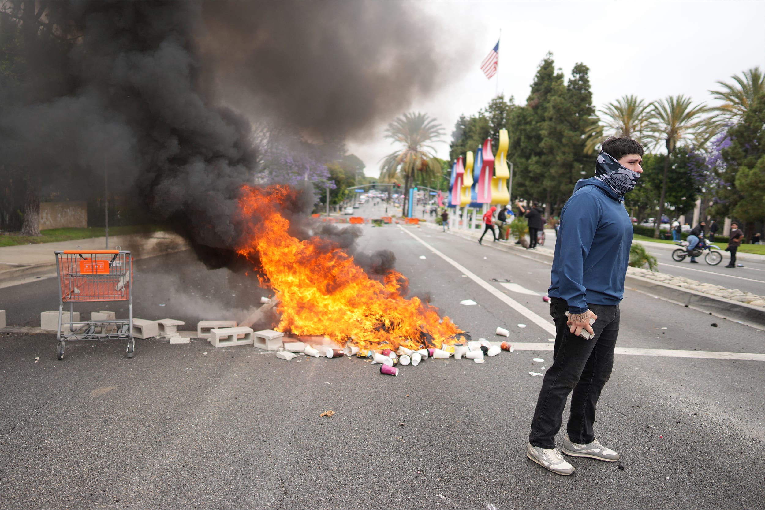 A fire burns as a protester stands across the way from Border Patrol personnel in riot gear and gas masks outside an industrial park in the city of Paramount, south of downtown Los Angeles, on Saturday, June 7, 2025.
