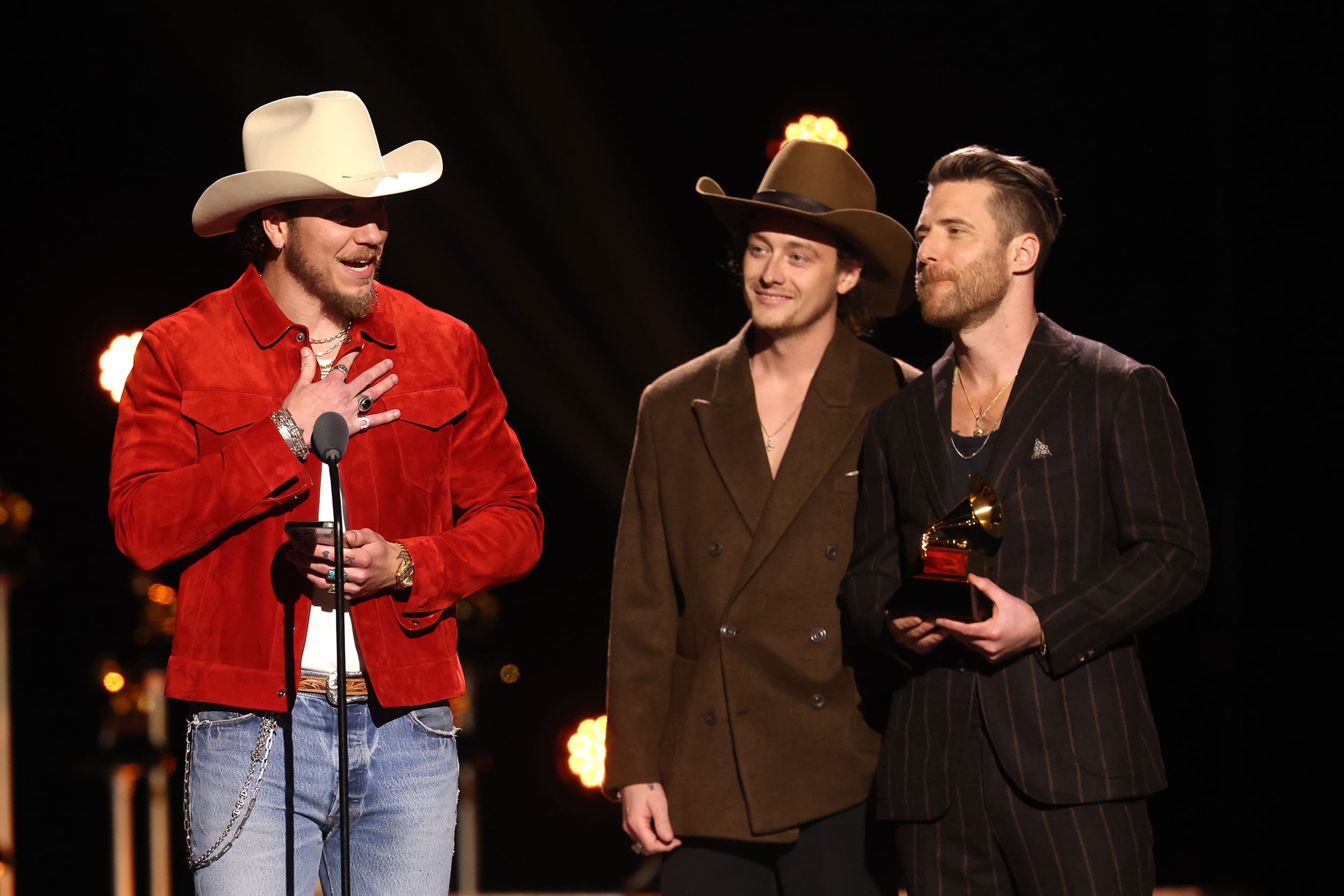 A man in a red jacket and cowboy hat holds a phone in one hand while speaking into a microphone, on stage during the Grammy awards.