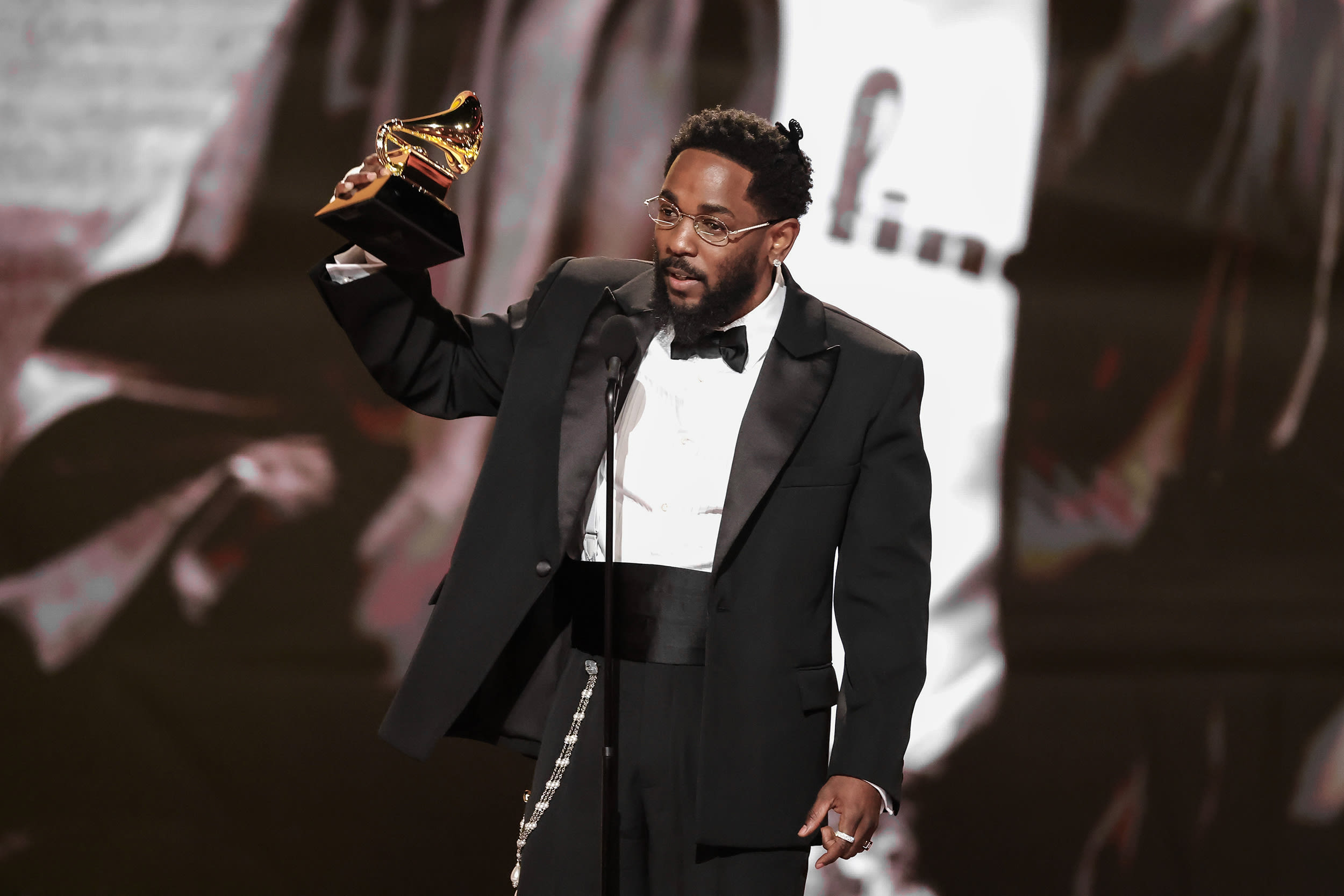 Kendrick Lamar holds a Grammy award in his right hand while standing at a microphone wearing a black tuxedo.