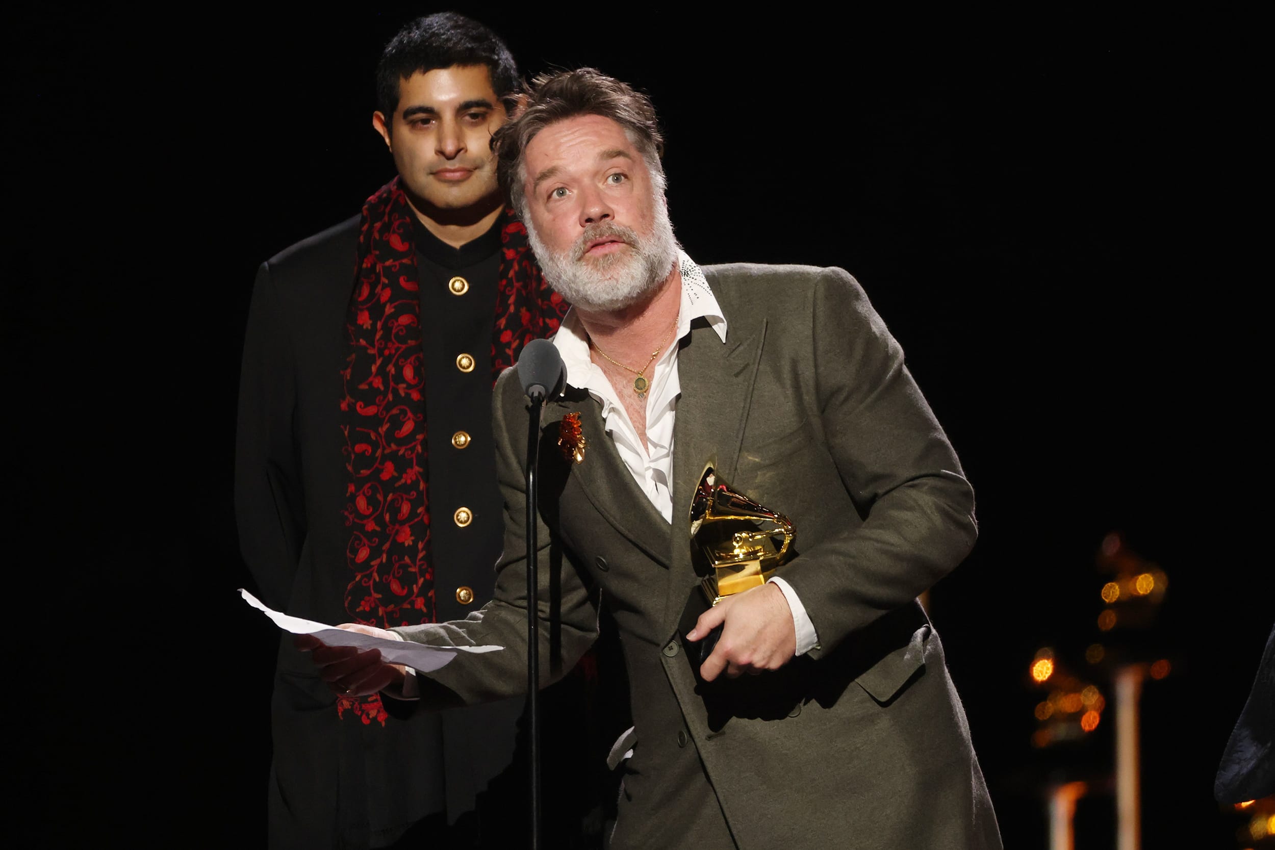 Rufus Wainwright holds a golden Grammy award in his left hand while leaning into a microphone, on stage at the Grammy awards.