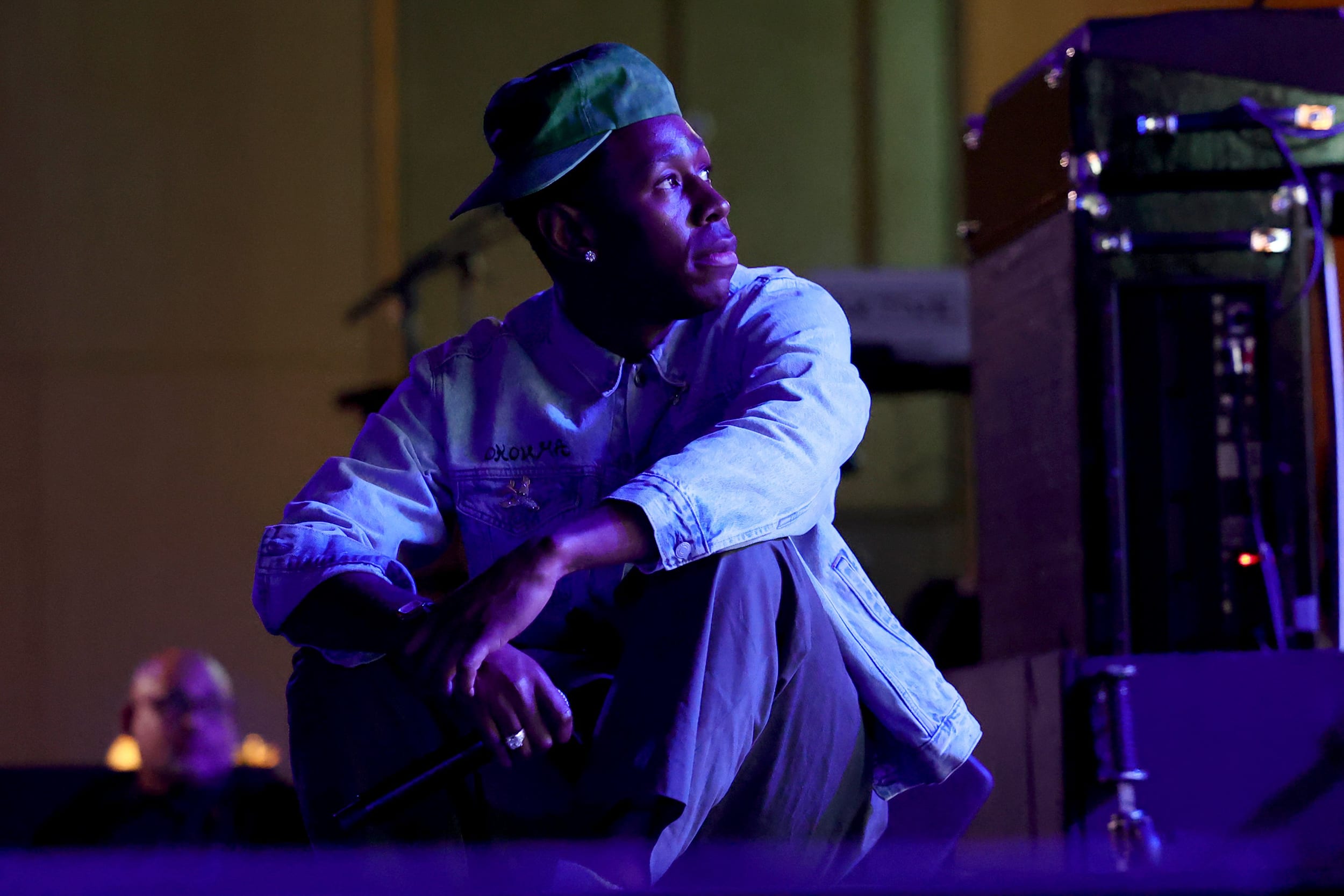 Tyler the Creator sits on a stage while holding a wireless microphone. He is illuminated by blue and purple stage lighting.