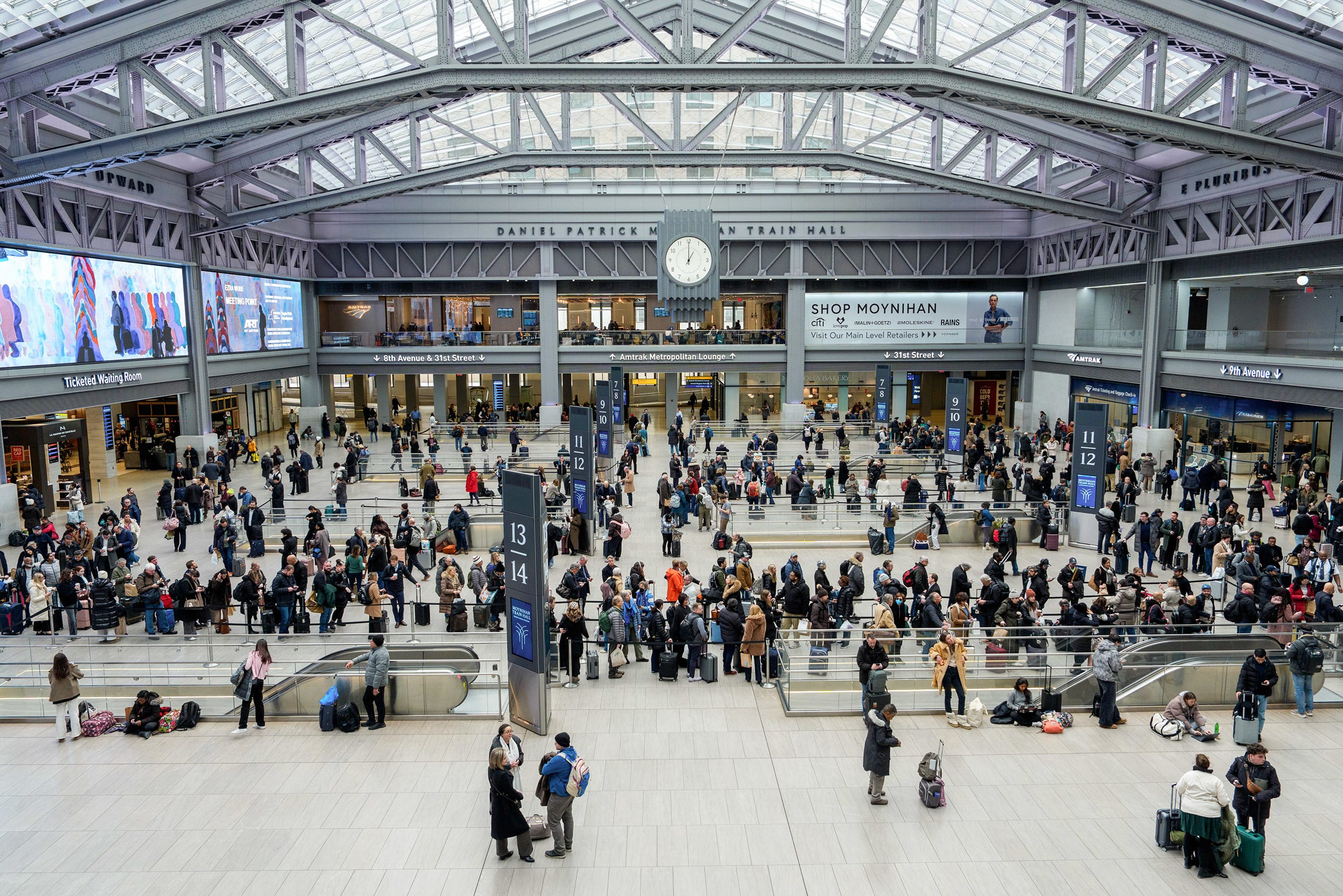 A crowd of people fills a large train hall with a glass roof, seen from an elevated perspective.