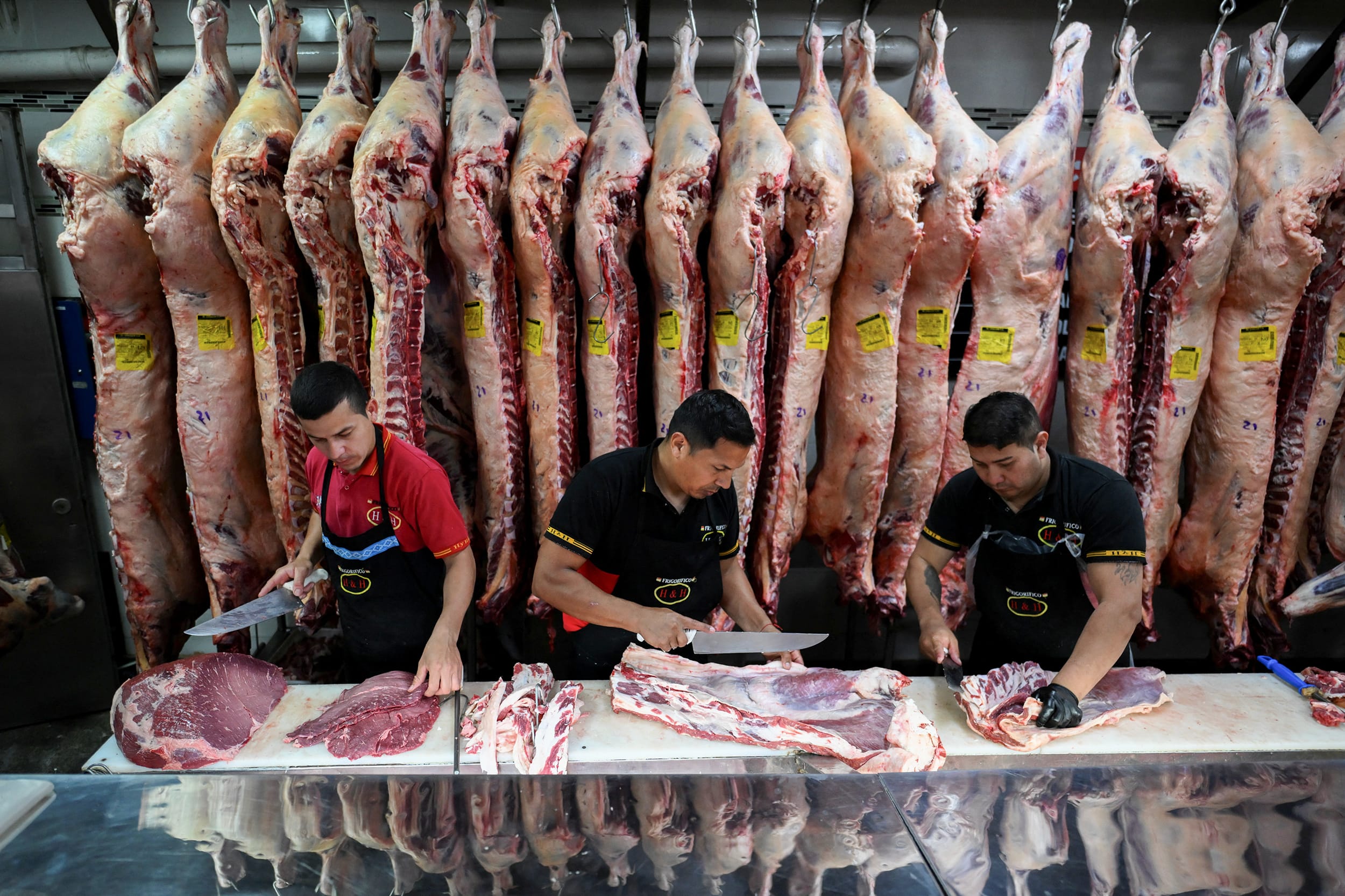 Three men wielding large butcher knives cut beef at a table in front of several large carcasses hanging from hooks.
