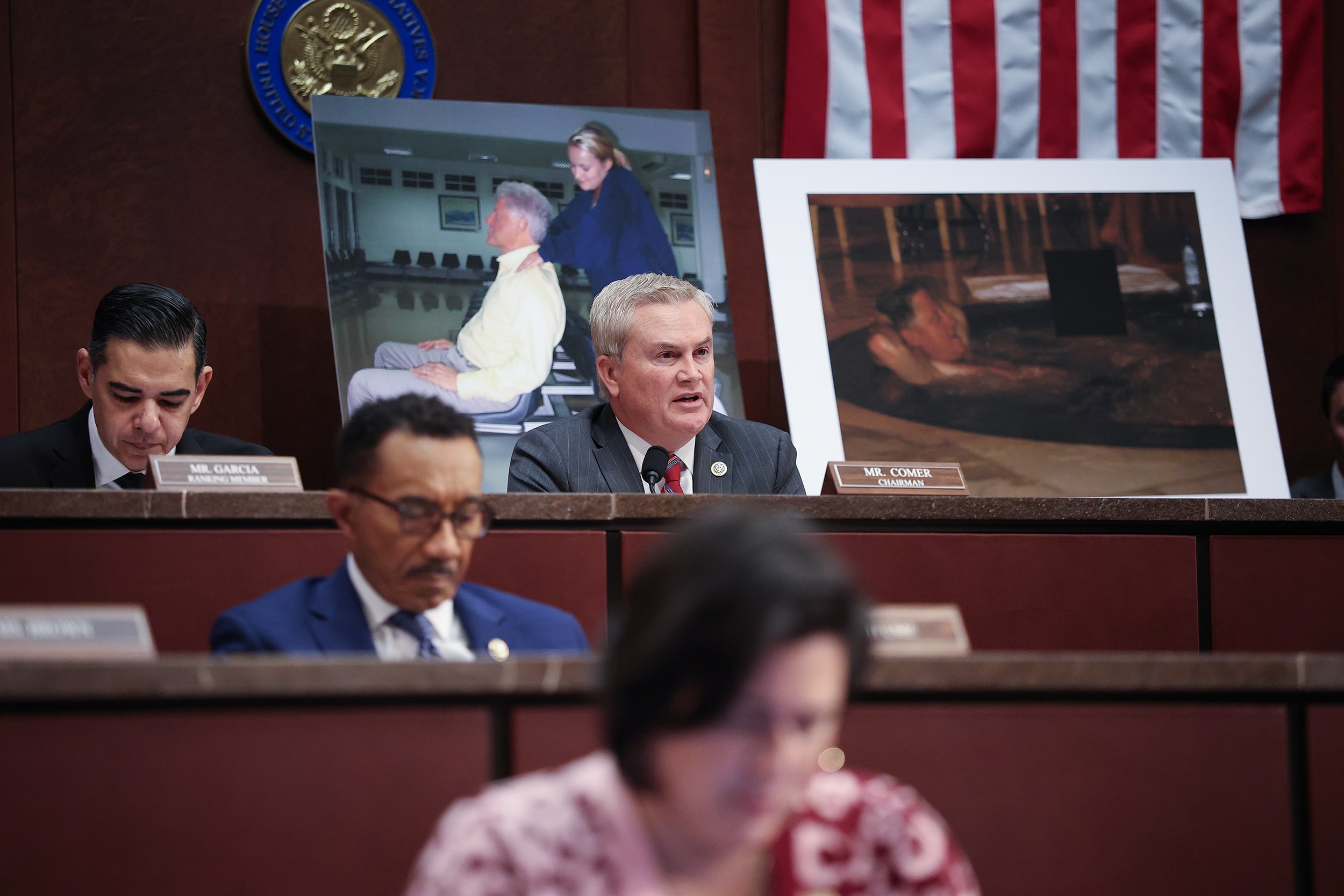 James Comer speaks while seated behind a microphone. A large print of former president Bill Clinton receiving a backrub from a blonde woman is displayed behind him.