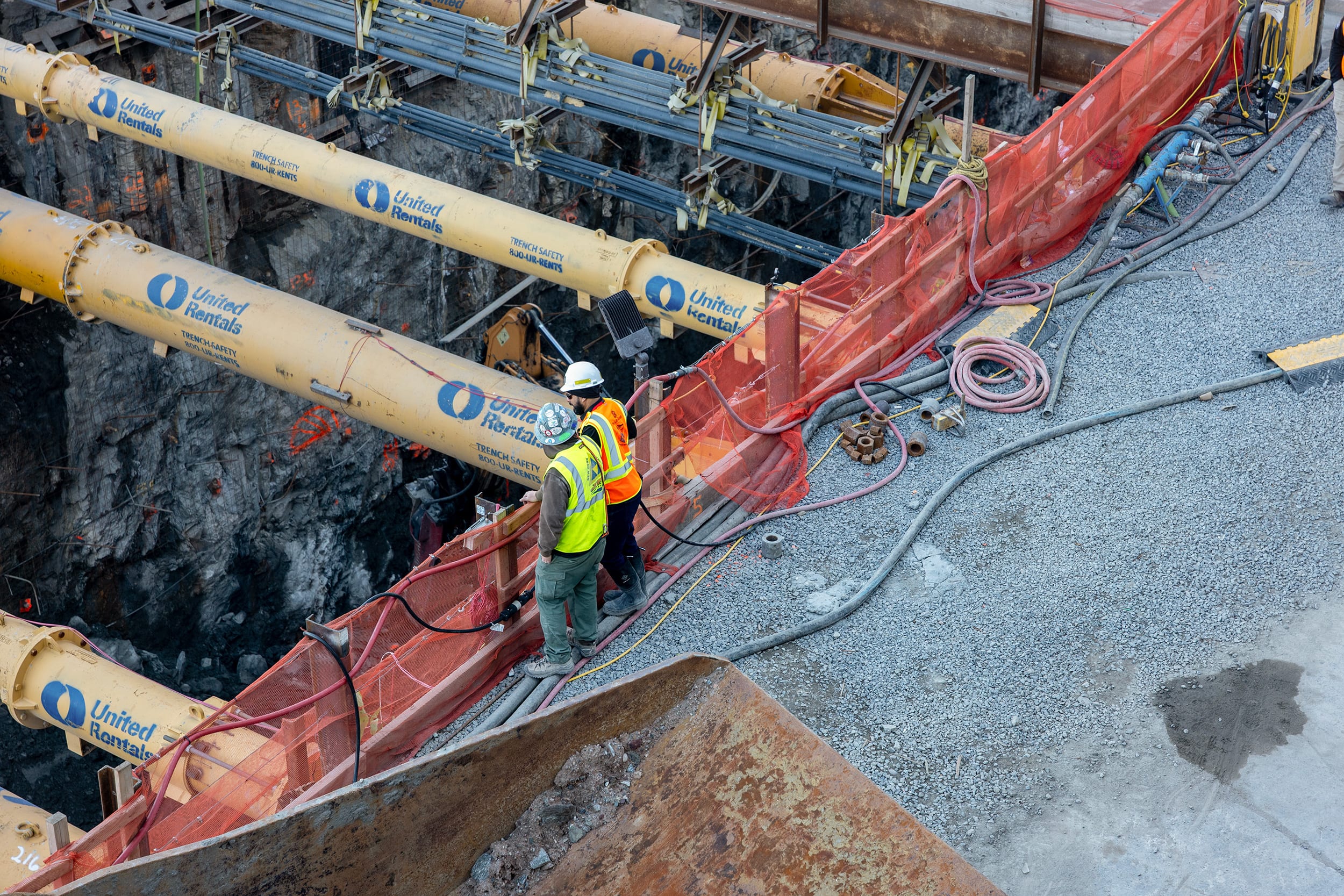 Two construction workers wearing neon safety vests and hardhats stand at the edge of an open pit with large pipes running across it.