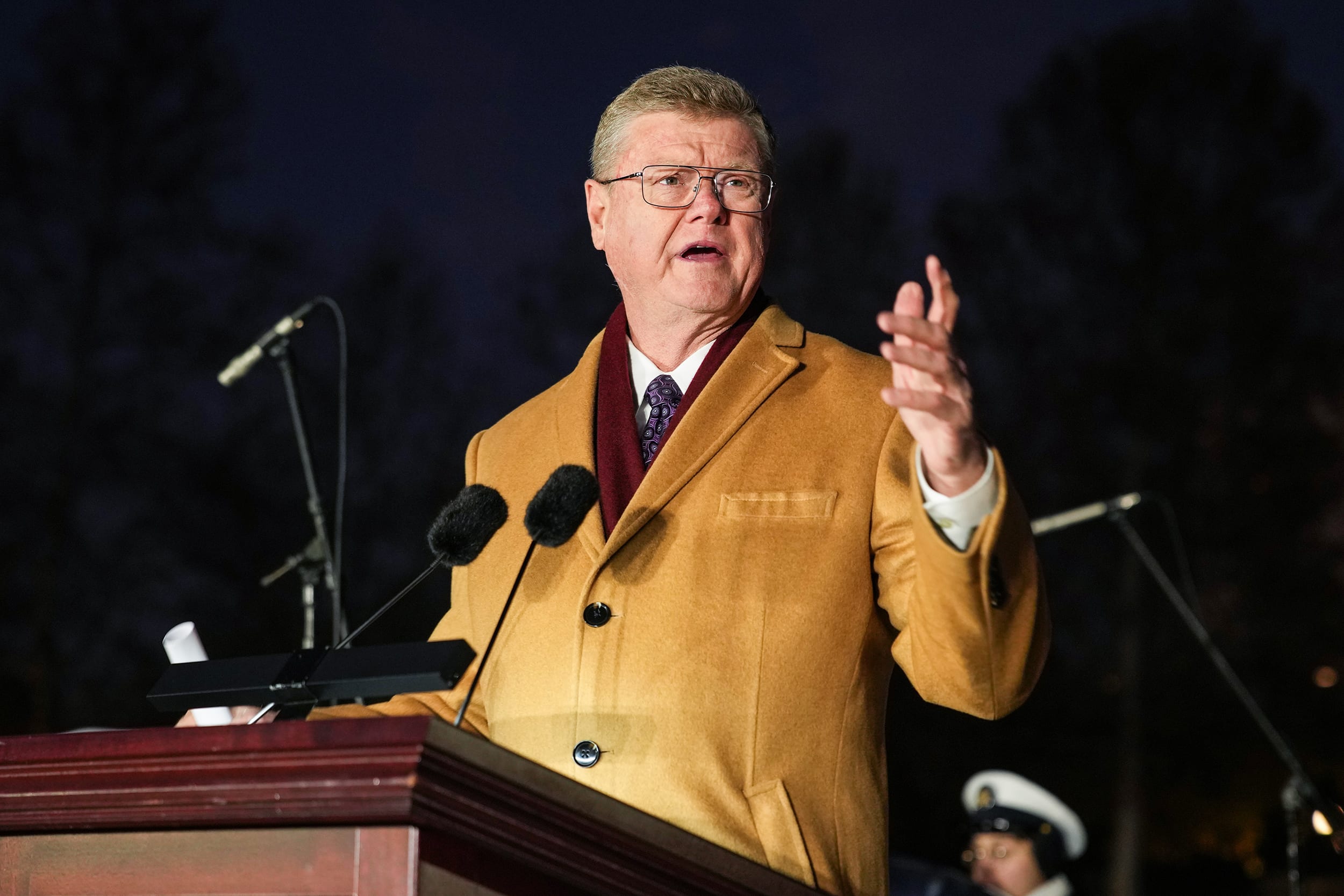 Mark Amodei speaking at a lectern during an outdoor event at night. He is wearing a tan wool coat.