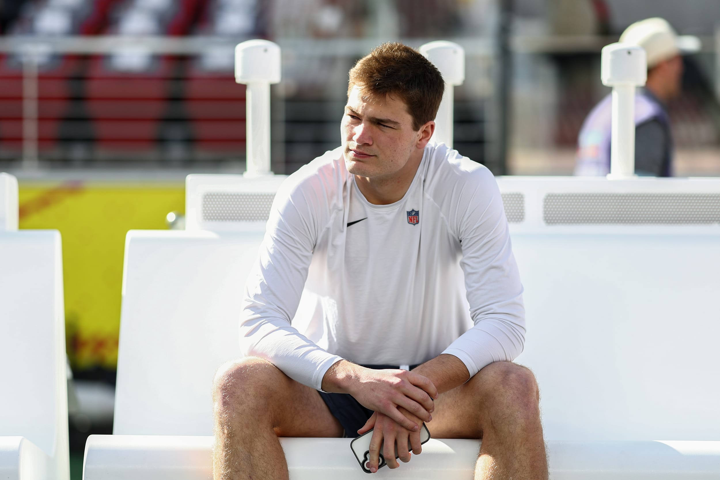 Drake Maye of the New England Patriots sits on a white bench on the sidelines. He's wearing a longsleeve white t-shirt and no football pads or helmet.