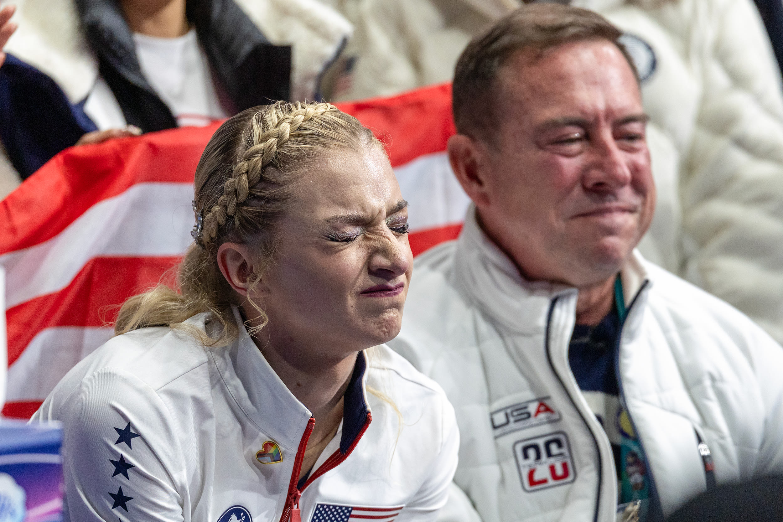 Amber Glenn grimaces with both eyes closed while sitting in front of an American flag.